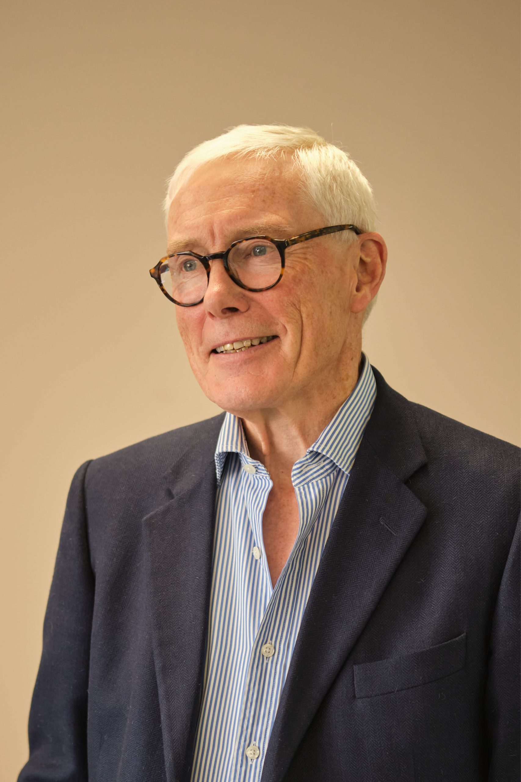 The image shows an older man wearing glasses, a navy blazer, and a blue and white striped shirt. The background is plain.