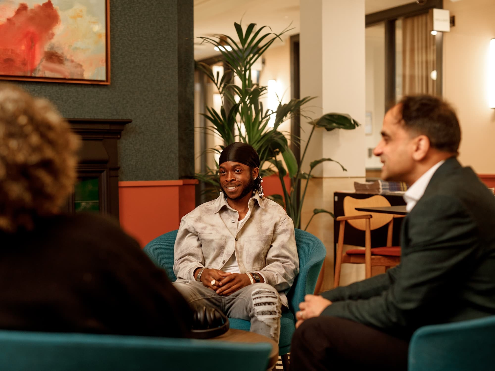 The image shows three people sitting in a room. One person is facing the camera and appears to be engaged in conversation with the other two, who are facing away. The setting includes modern furniture, plants, and a painting on the wall, giving it