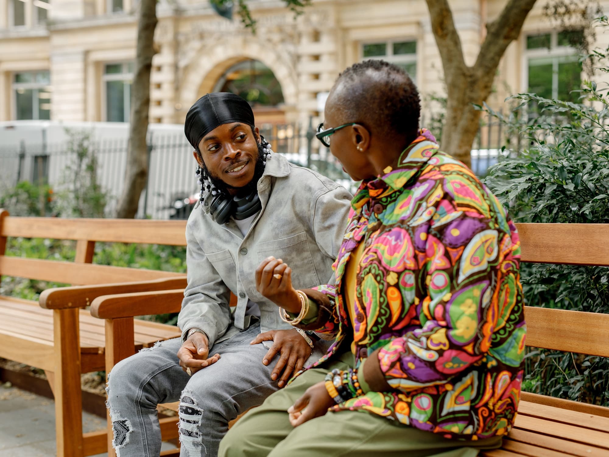 The image shows two people sitting on a bench outdoors, engaged in conversation. One person is wearing a light-colored jacket, headphones around their neck, and a dark head wrap. The other is wearing a brightly patterned shirt and glasses. They appear to be