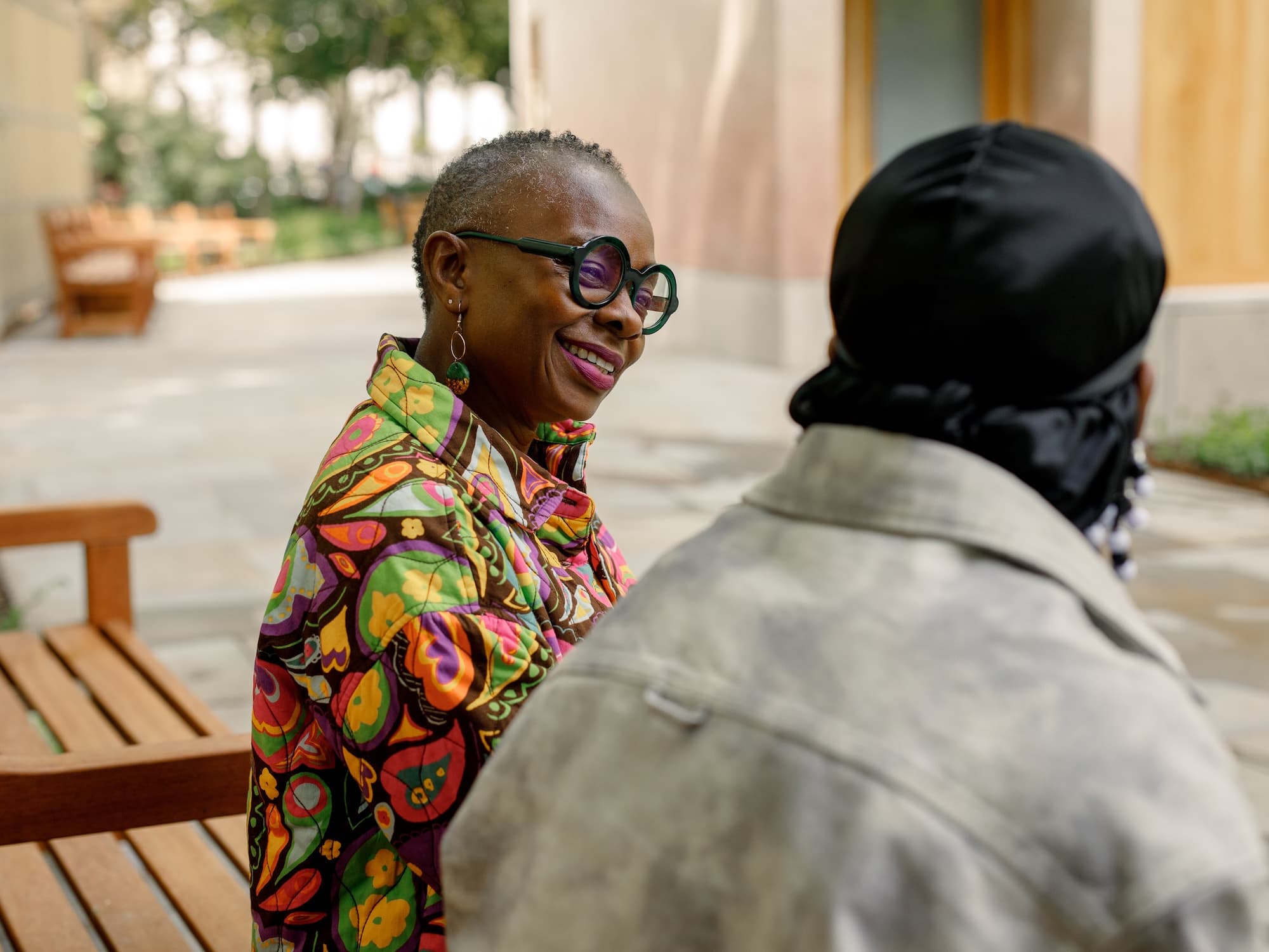 The image shows two people sitting on a wooden bench outdoors. One person is wearing a colorful, patterned jacket and round glasses, and is smiling at the person across from them, who is wearing a dark head covering and light-colored jacket. The background features