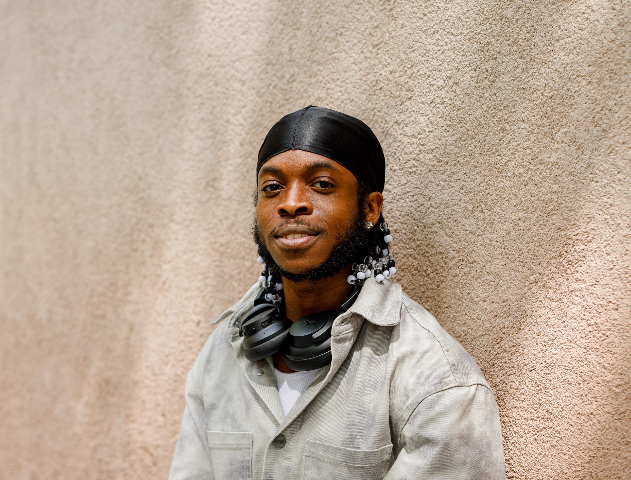 The image shows a person wearing a gray jacket and a black headscarf. They have beaded hair and are wearing headphones around their neck, smiling and standing against a textured wall.