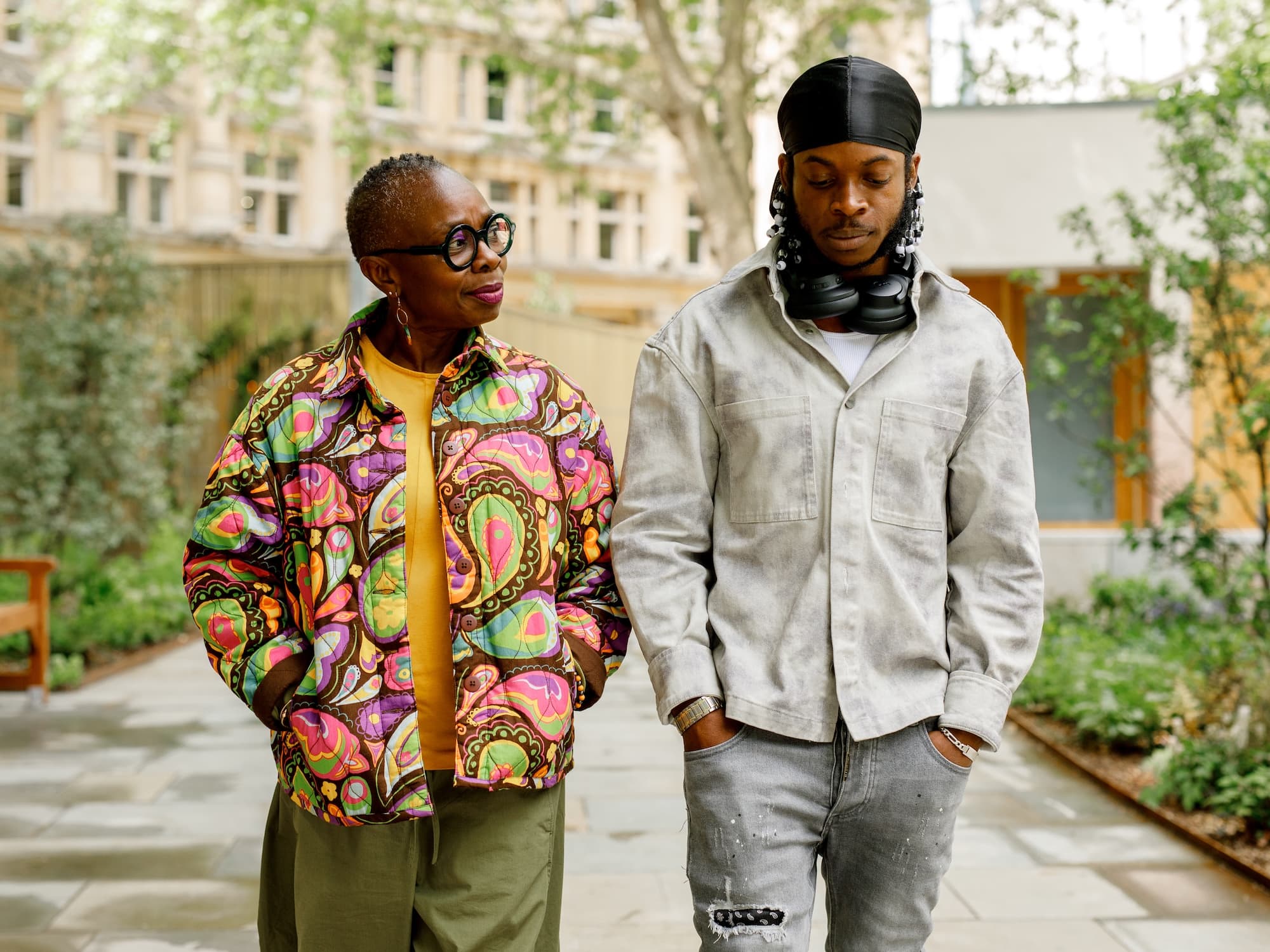 The image shows two people walking outdoors. The person on the left is wearing a brightly colored, patterned jacket and glasses, while the person on the right is wearing a light-colored shirt, a black head wrap, and headphones around their neck. They appear