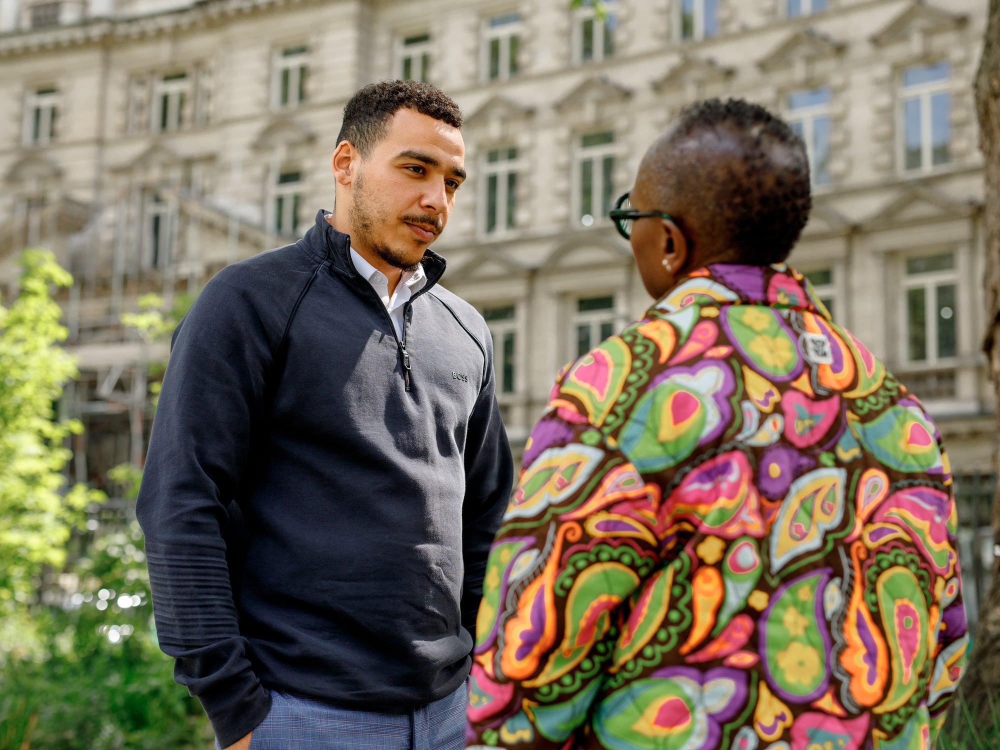 The image shows two people standing outside in front of a building with ornate architecture. One person is wearing a dark pullover, while the other is wearing a colorful, patterned jacket. They appear to be engaged in conversation. There are trees and greenery around