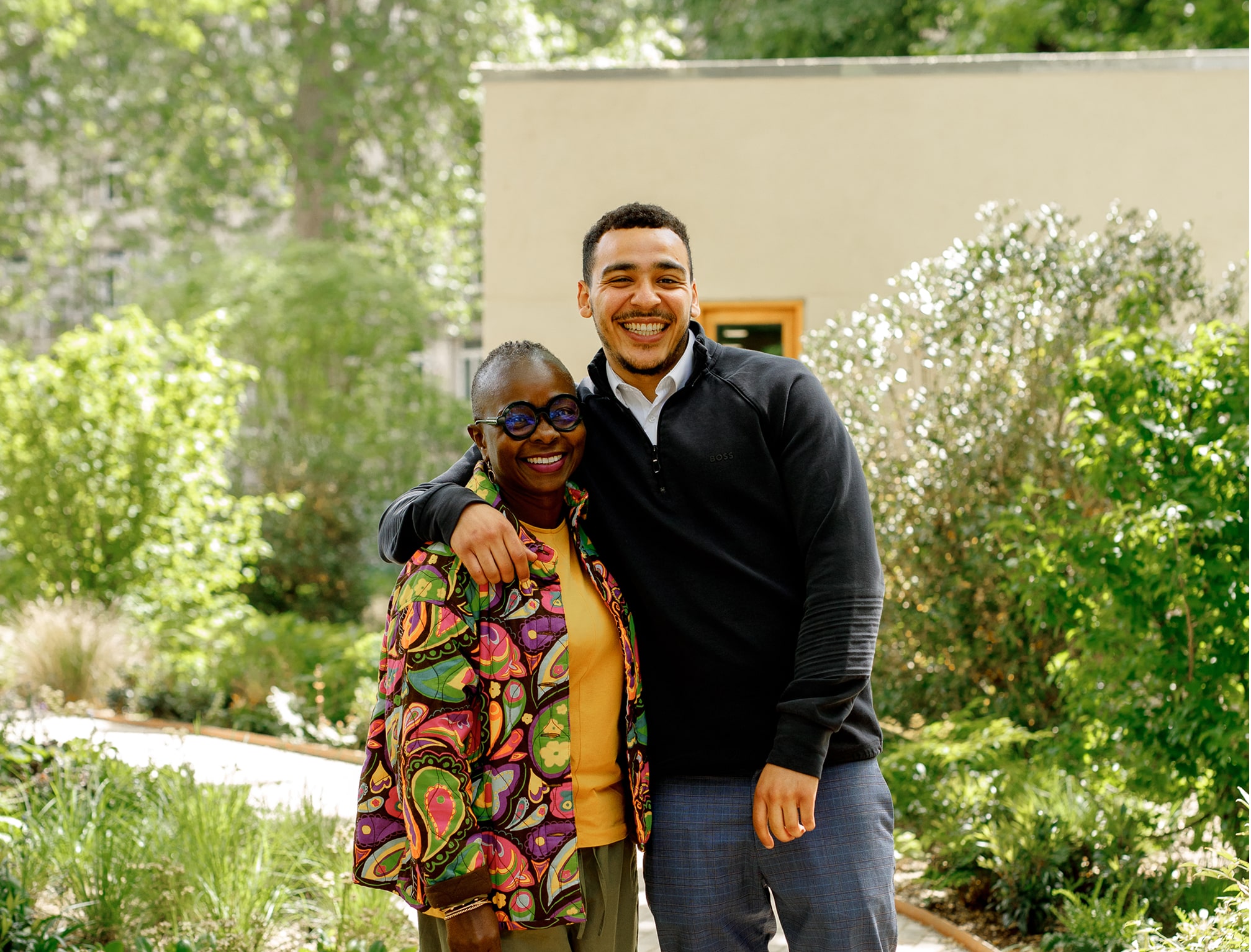 The image shows two people standing outdoors in a garden-like setting. They are smiling and appear to be posing for the photo, with one person having their arm around the other. The background features greenery, including trees and shrubs.