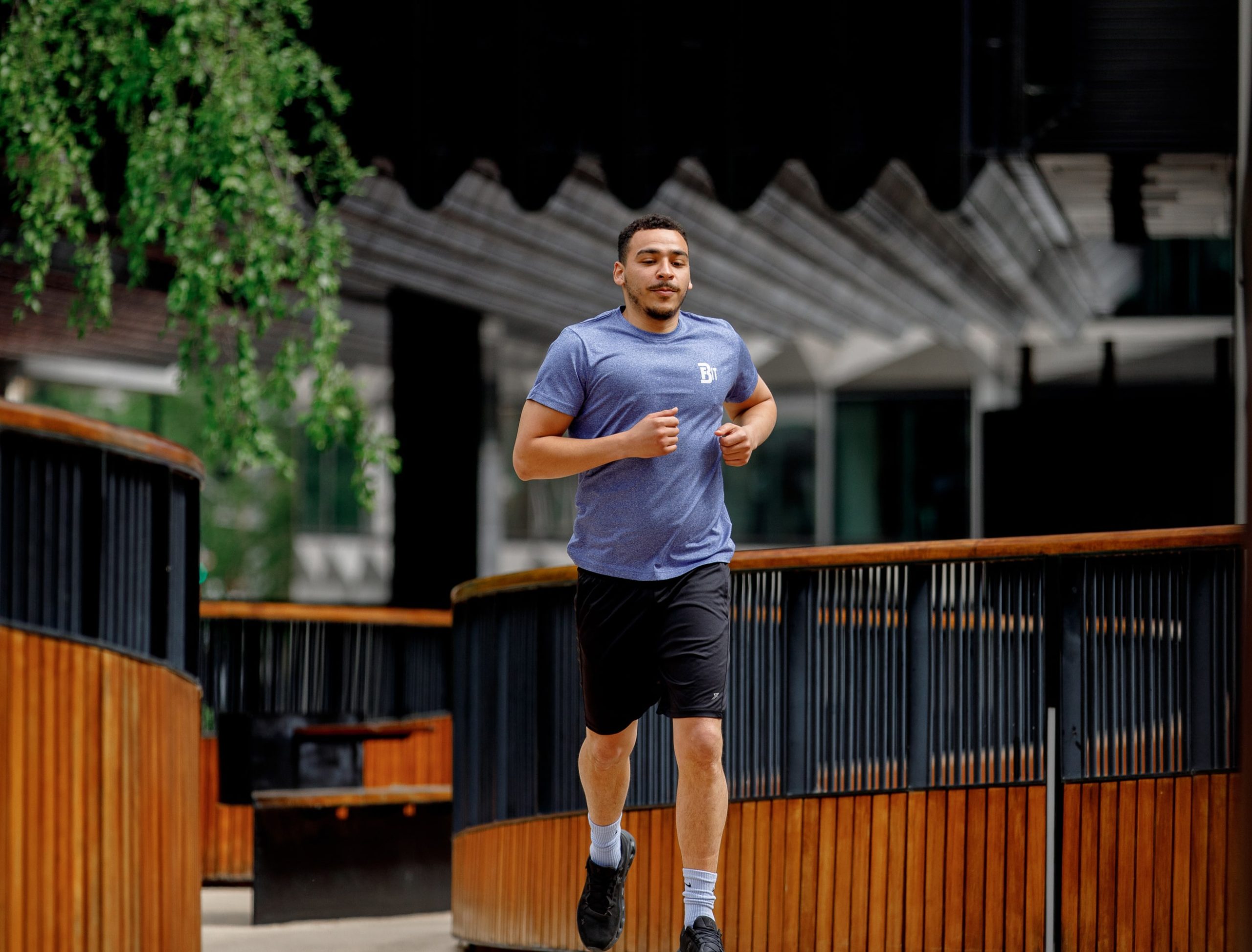 The image shows a person jogging outdoors. The individual is wearing a blue shirt and black shorts and appears to be on a wooden boardwalk or path with metal railings. There are trees and a modern building in the background.