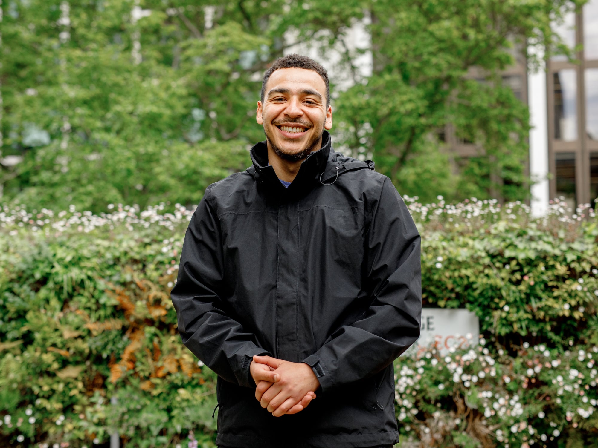 The image shows a person standing outdoors, smiling, and wearing a black jacket. The background is filled with greenery, including bushes and trees, suggesting a garden or park setting.