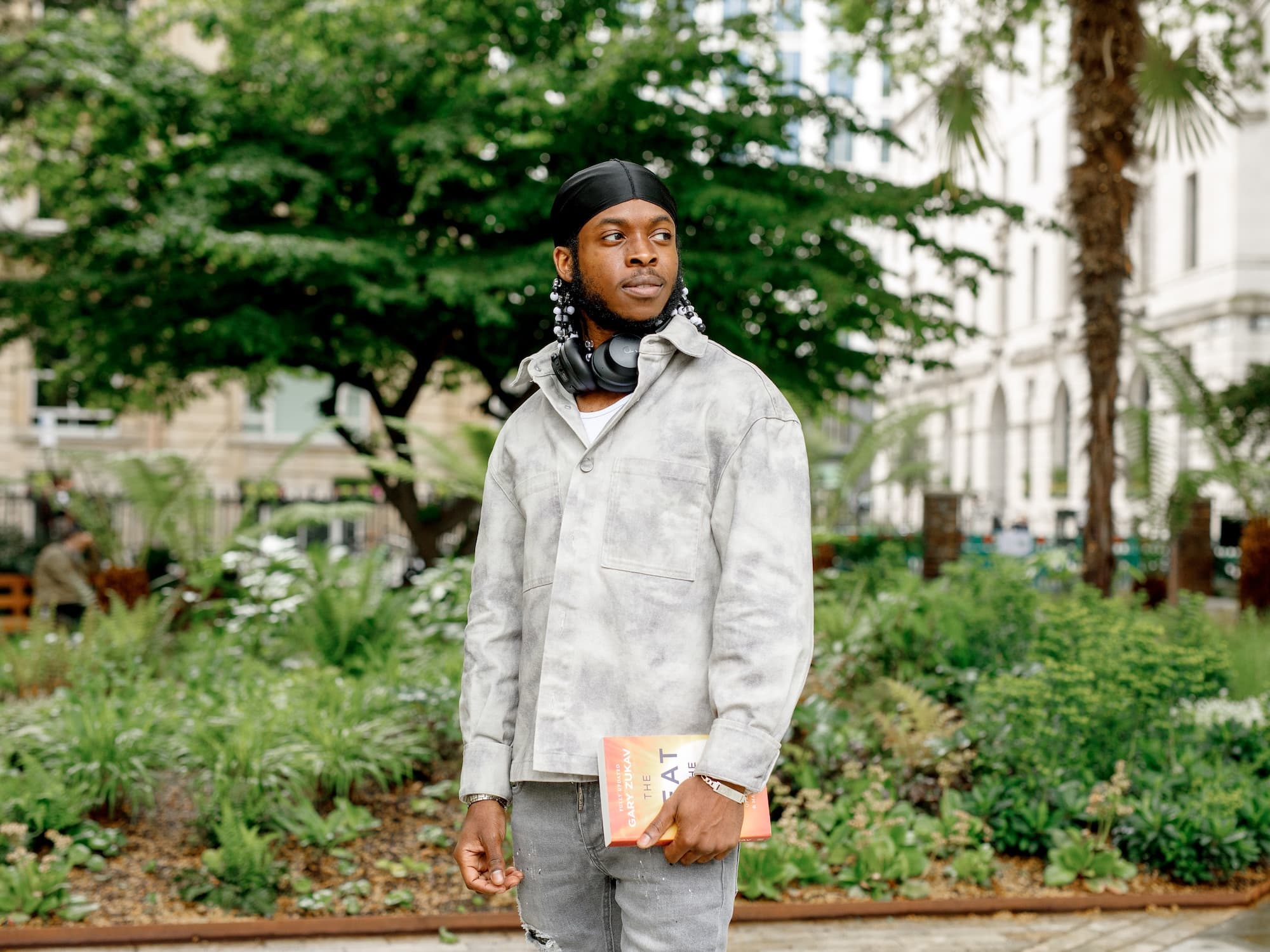 The image depicts a person standing outdoors in a garden-like setting. They are wearing a light-colored jacket and dark headwear, with headphones around their neck. The person appears to be holding a book. The background consists of greenery and some buildings.