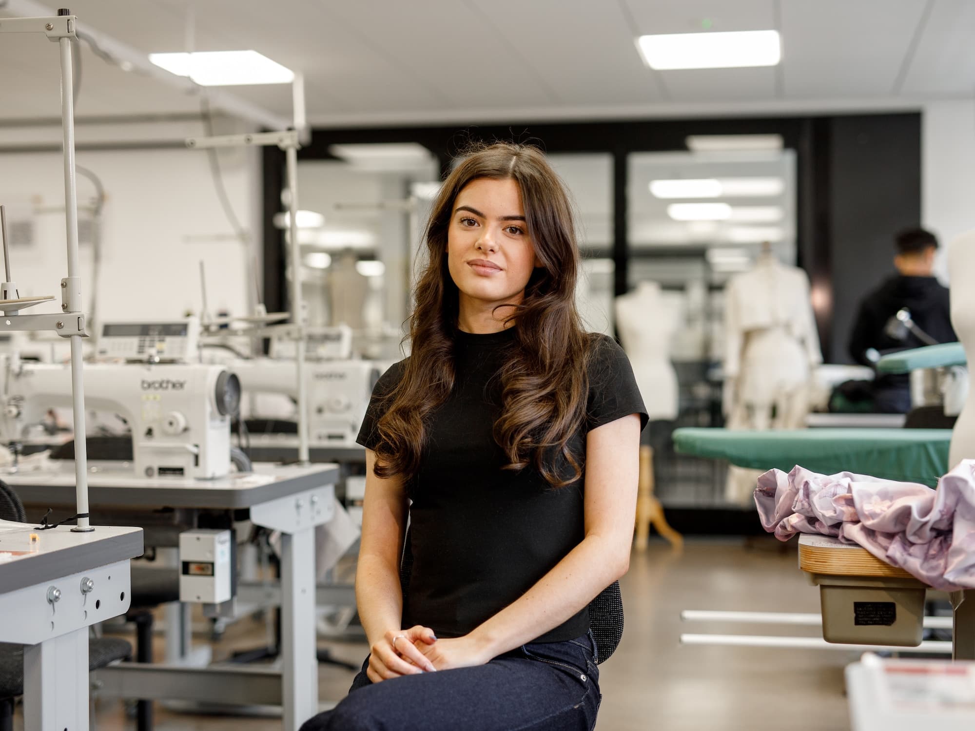 The image shows a person sitting in a sewing or fashion design studio. There are several sewing machines in the background and some fabric on a table. The environment has a clean, professional appearance and is likely a workspace for fashion-related activities.