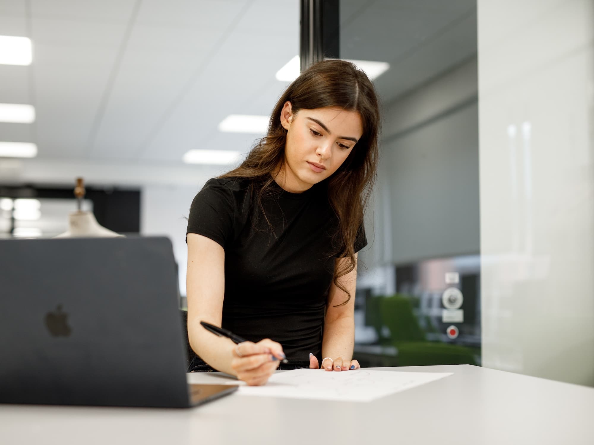 The image shows a person sitting at a desk, writing or drawing on paper. A laptop is open on the desk, and the setting appears to be an office or classroom with a mannequin in the background. The person is focused on their work.