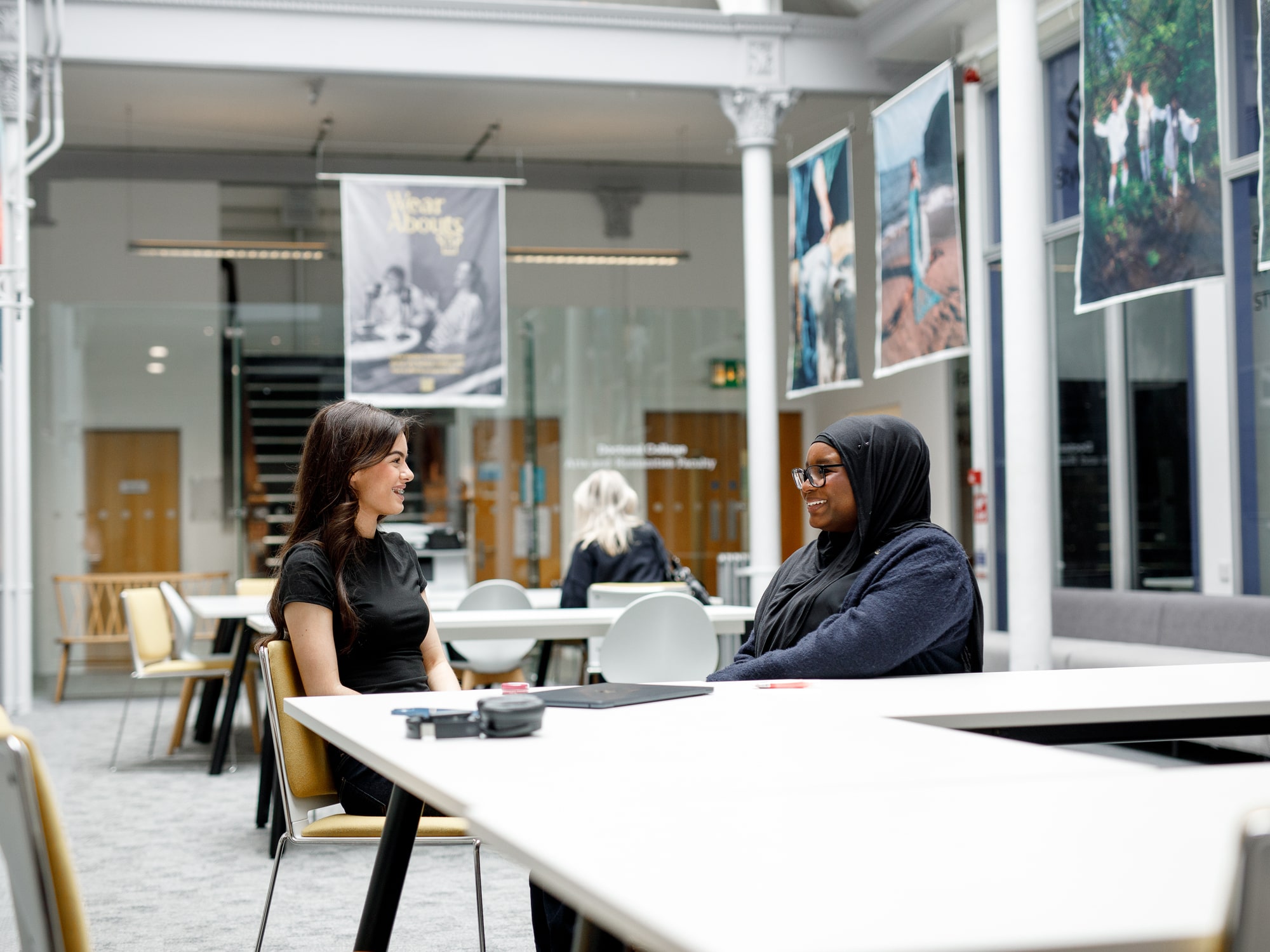 This image shows two people sitting at a table in a modern, open indoor setting, possibly a library or a co-working space. They appear to be engaged in conversation. There are posters hanging from the ceiling and the space has a bright, airy feel