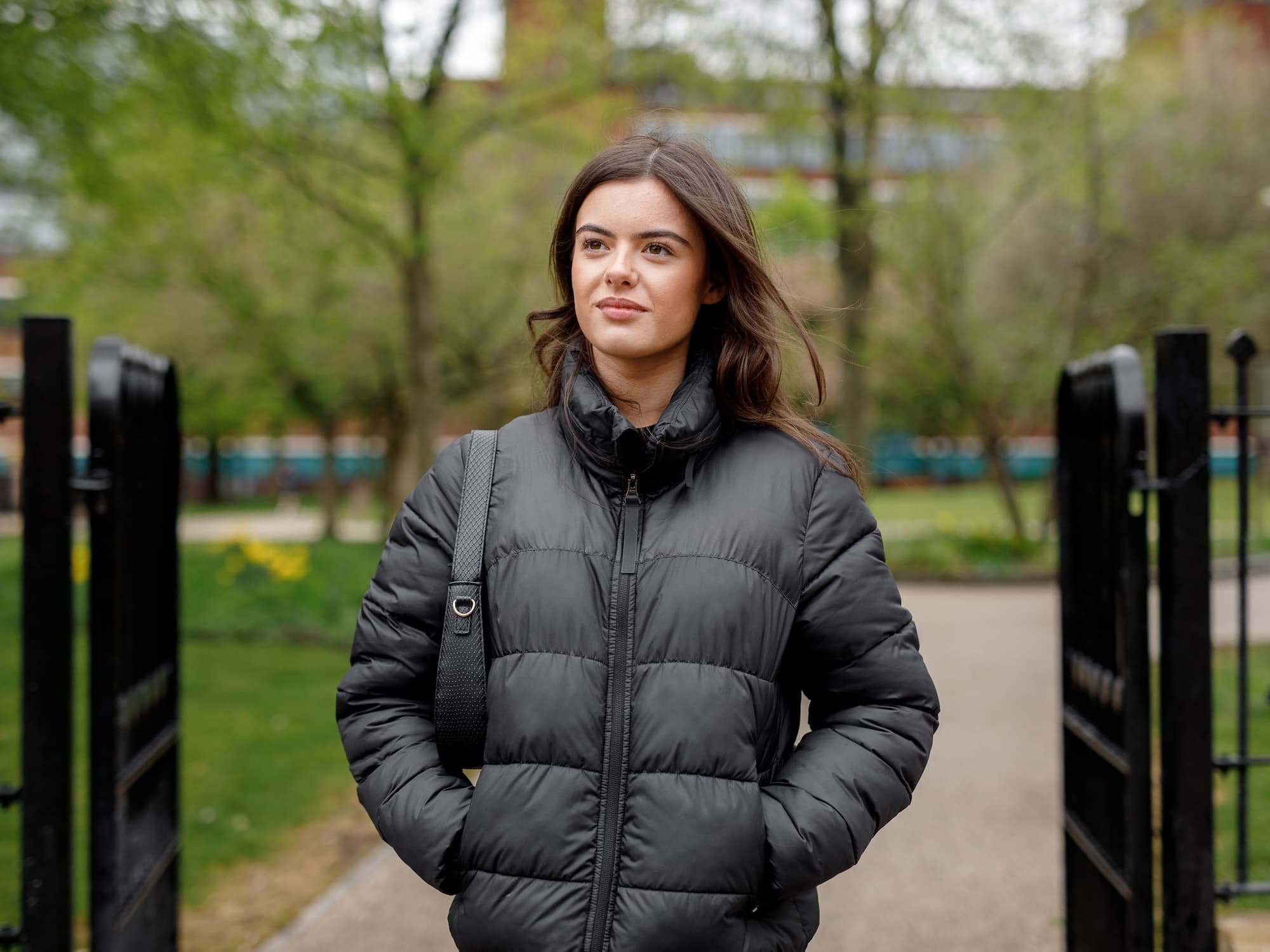 The image shows a person wearing a black puffer jacket, standing outdoors. The background includes trees and a pathway, suggesting a park or garden setting. The person is looking slightly to the side with hands in pockets.