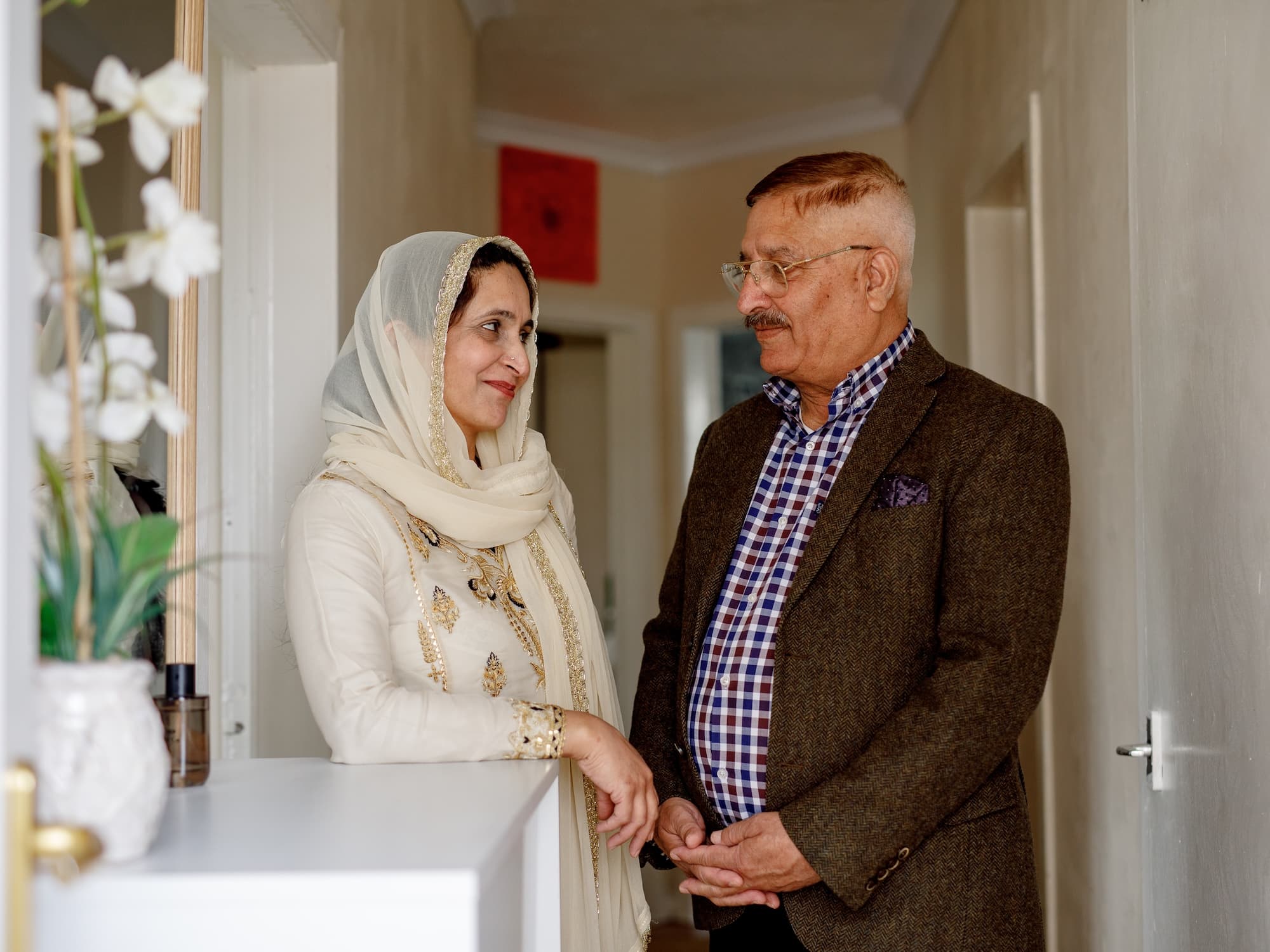 The image shows a man and a woman standing in a hallway, engaging in conversation. The woman is wearing a light-colored, embroidered outfit with a matching headscarf, while the man is dressed in a checked shirt and a brown blazer. They appear