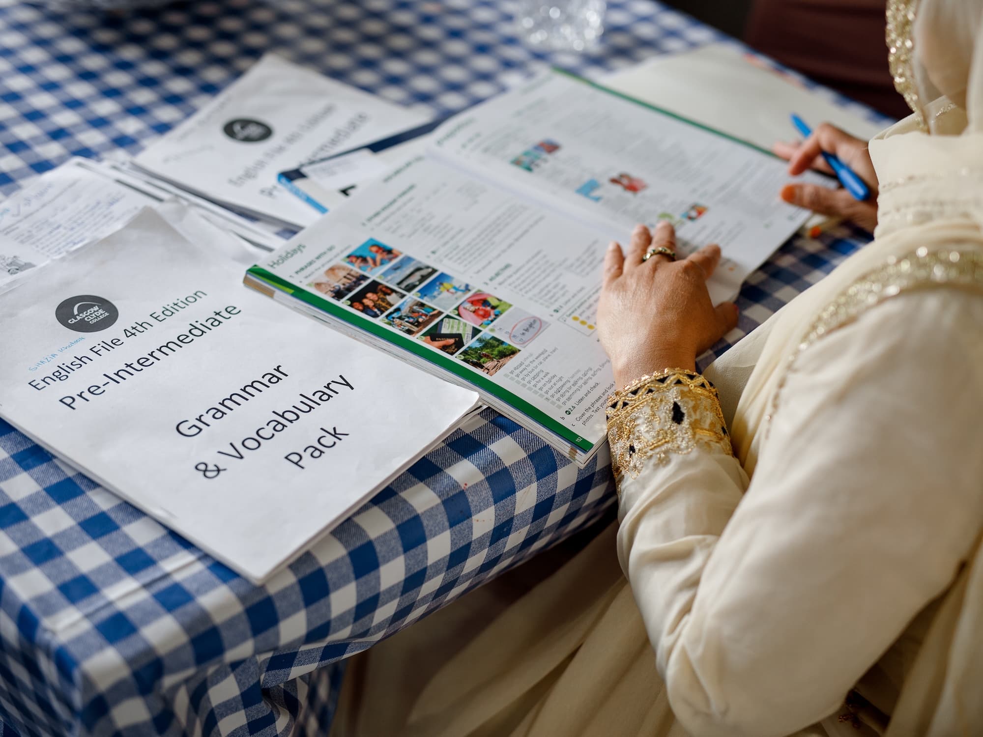 The image shows a person reading a book or workbook related to language learning on a blue and white checkered tablecloth. There are several papers, including one titled English File 4th Edition Pre-Intermediate Grammar & Vocabulary Pack. The person is