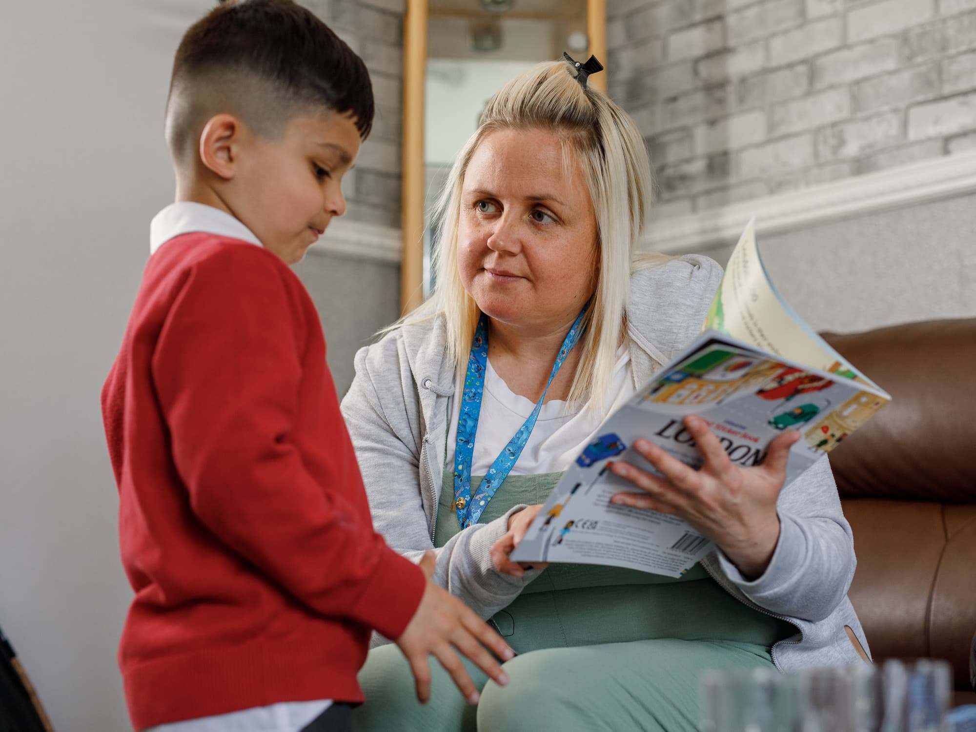 The image shows a woman sitting and holding a book open, while looking at a young boy standing beside her. The boy is wearing a red sweater and appears to be engaging with the woman, who seems to be reading or discussing the book with him.