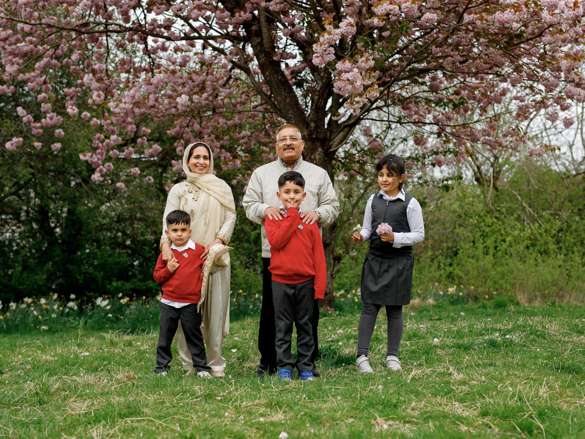 The image shows a family, including two adults and three children, standing on grass in front of a tree with blooming pink flowers. The children are wearing school uniforms, and the adults are dressed in traditional clothing. The scene appears to be a family portrait