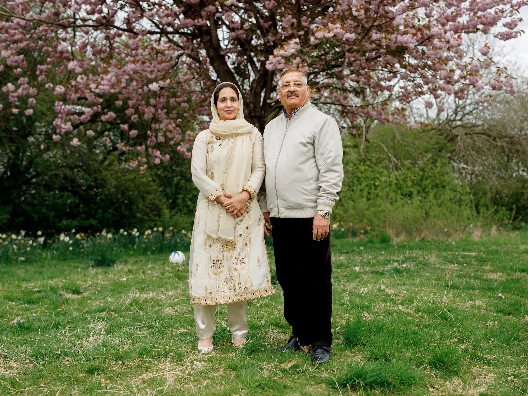 The image shows a man and a woman standing in a grassy area with a tree full of pink blossoms in the background. The woman is wearing a cream-colored outfit, and the man is in a light-colored jacket and dark pants. It appears to be