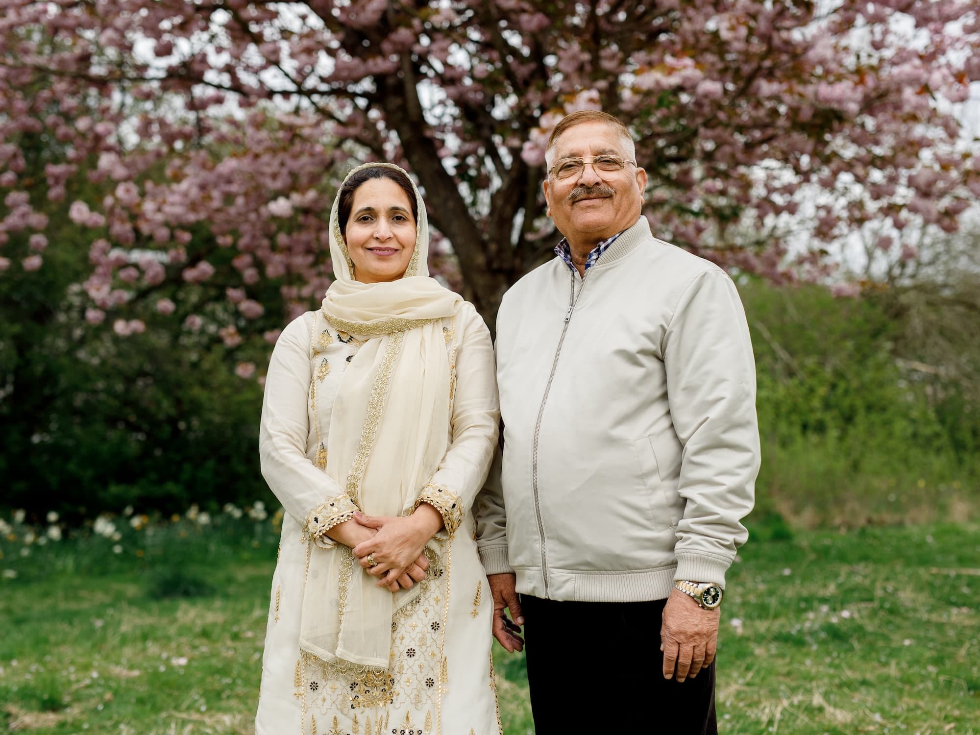The image shows two people standing outdoors in a grassy area. They are in front of a tree with pink blossoms. The woman on the left is wearing a traditional outfit with a headscarf, and the man on the right is wearing a light-colored
