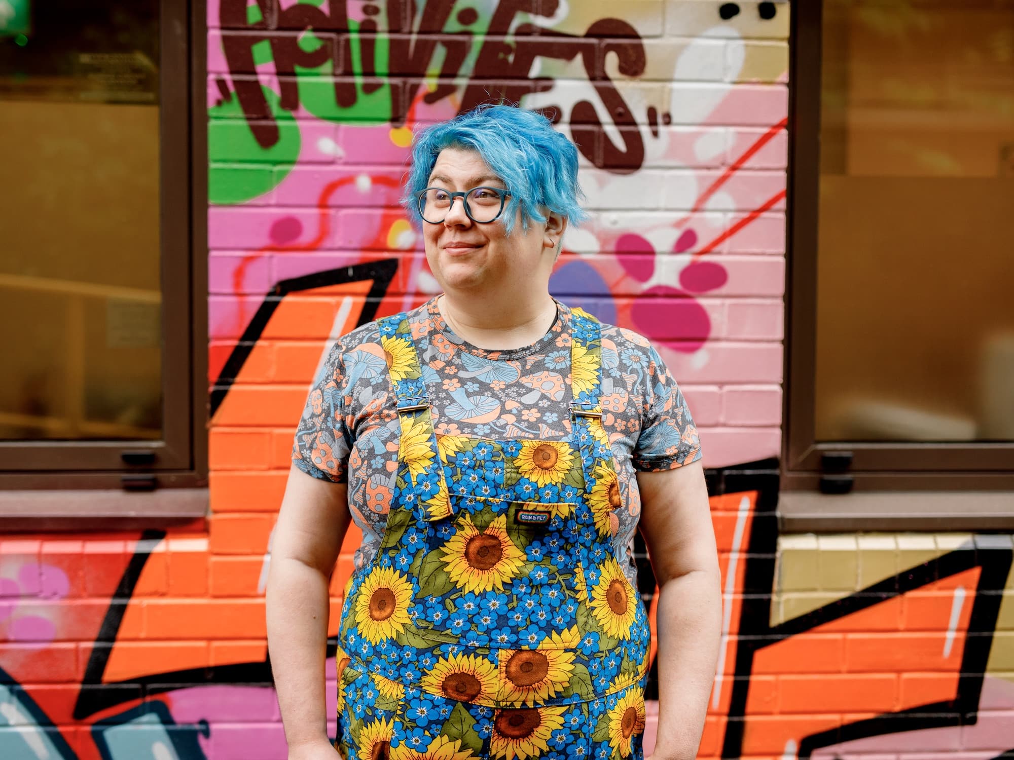 The image shows a person with blue hair standing in front of a vibrant graffiti-covered wall. They are wearing glasses and colorful clothing featuring a pattern of sunflowers and other floral designs. The wall behind them is painted with bright colors and abstract designs.