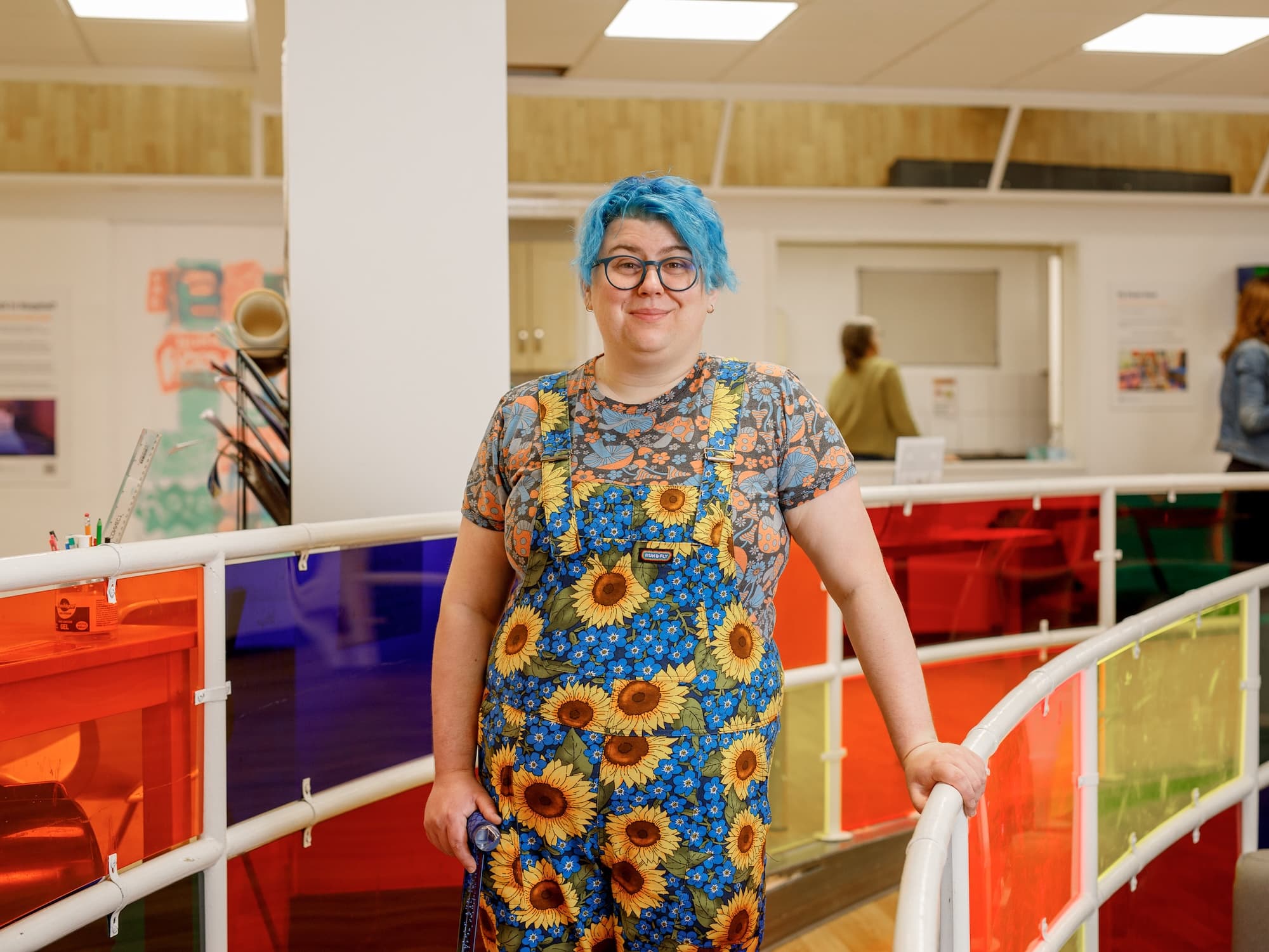 The image shows a person with blue hair and glasses standing in an indoor setting. They are wearing a floral-patterned outfit with sunflowers and blue flowers. The background features colorful panels on a railing and some people in the distance, suggesting a casual or