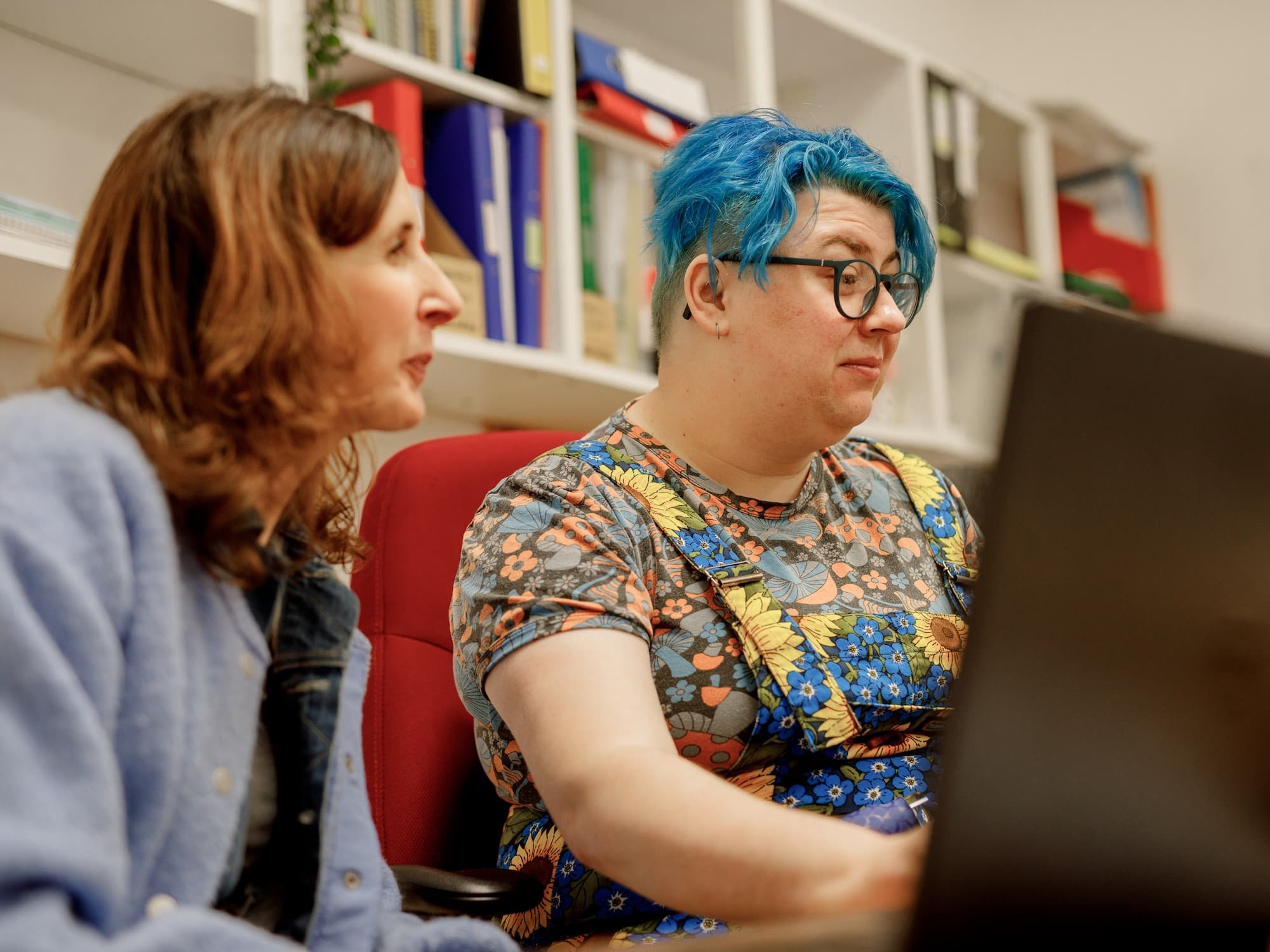 The image shows two people in a room with shelves in the background. One person has blue hair and is wearing glasses and a colorful floral outfit, while the other person has long brown hair and is wearing a light blue jacket. They appear to be focused