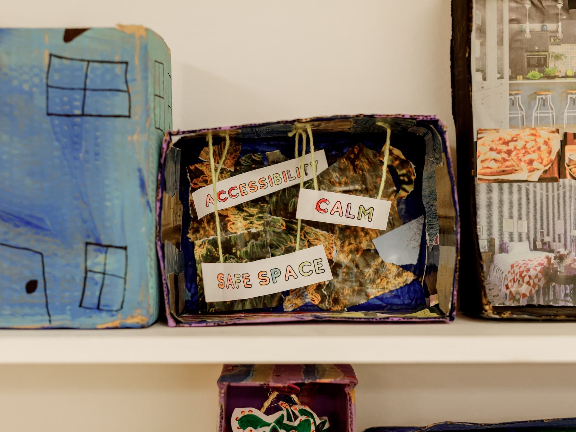 The image shows a shelf with a box containing cutouts and labels. The labels read ACCESSIBILITY, CALM, and SAFE SPACE. The box is part of a creative display, perhaps an art or school project. The surrounding boxes have