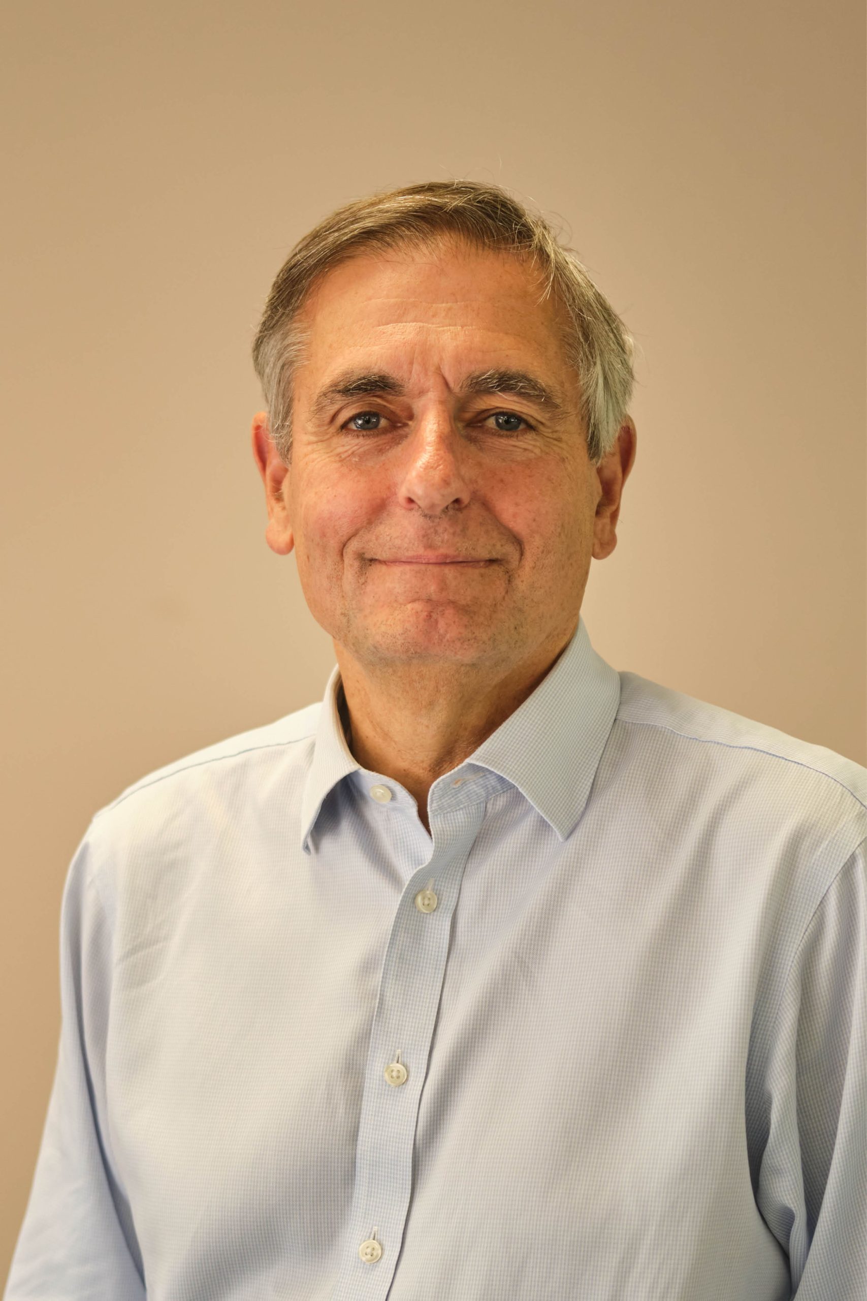The image shows an older man wearing a light blue button-up shirt, standing against a plain background.
