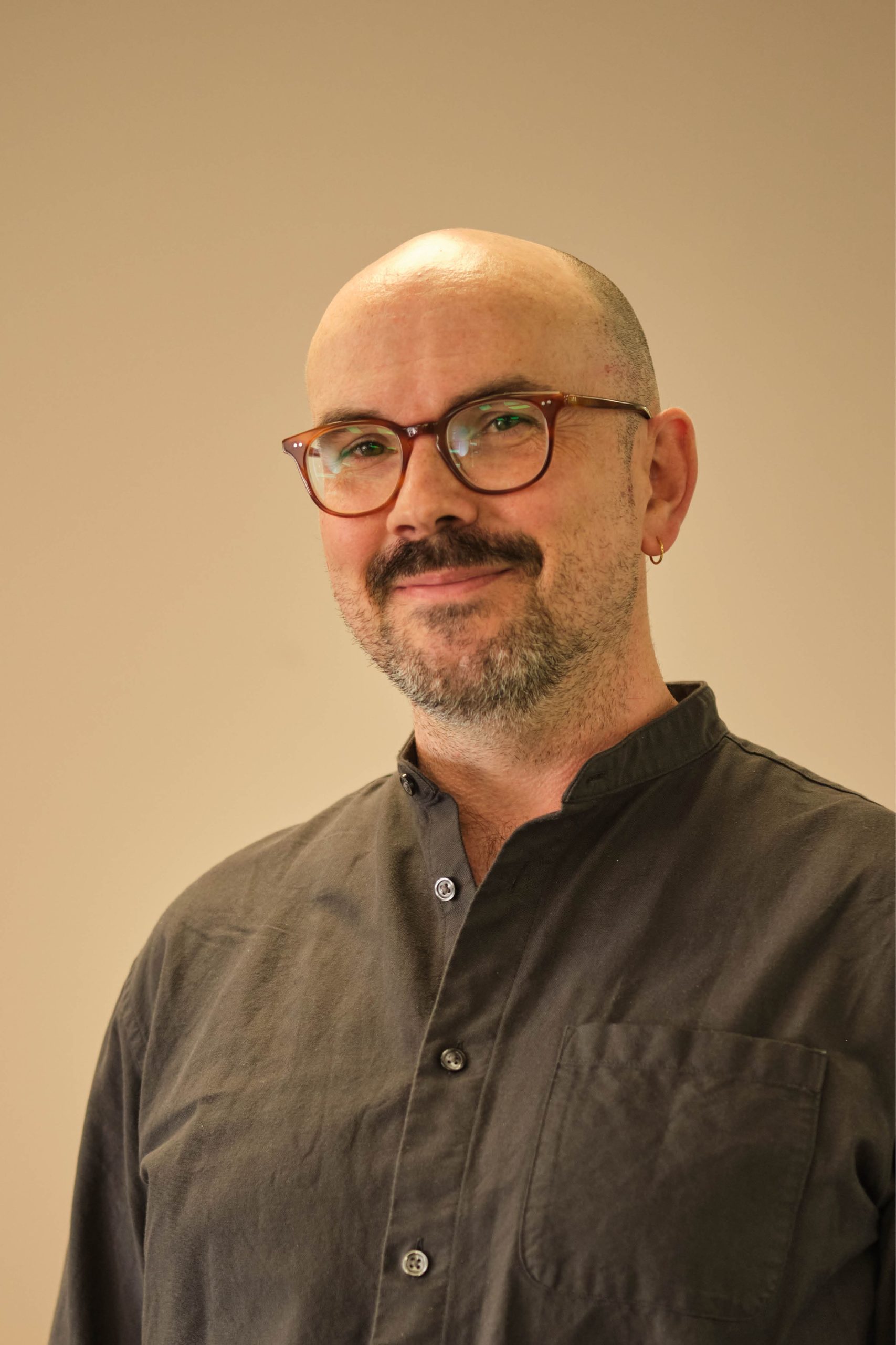 The image shows a bald man with glasses and facial hair, wearing a dark, buttoned-up shirt with a pocket. He has an earring in his left ear and is standing against a plain background.