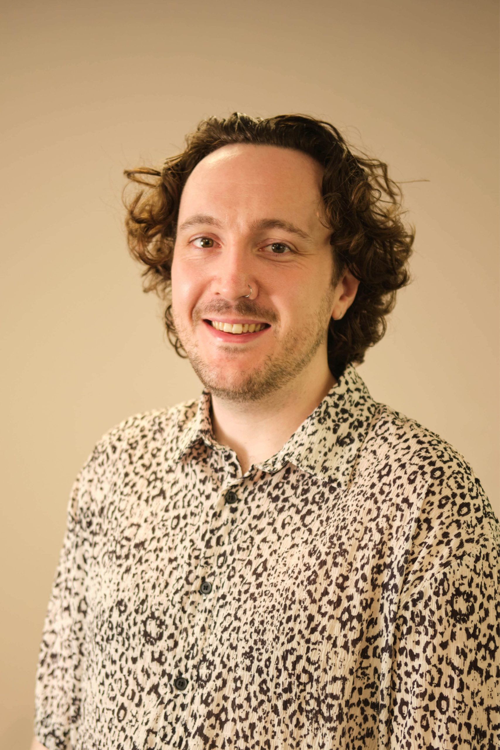 The image shows a person with curly hair smiling at the camera. They are wearing a leopard print shirt.