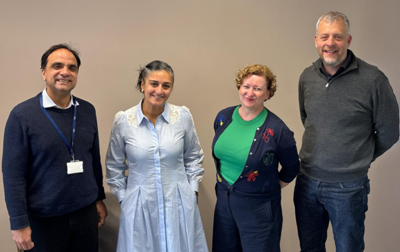 The image shows four people standing side by side against a plain background. They are dressed in casual to semi-formal attire. The person on the left is wearing a dark sweater and a lanyard with a badge. The other three are dressed in