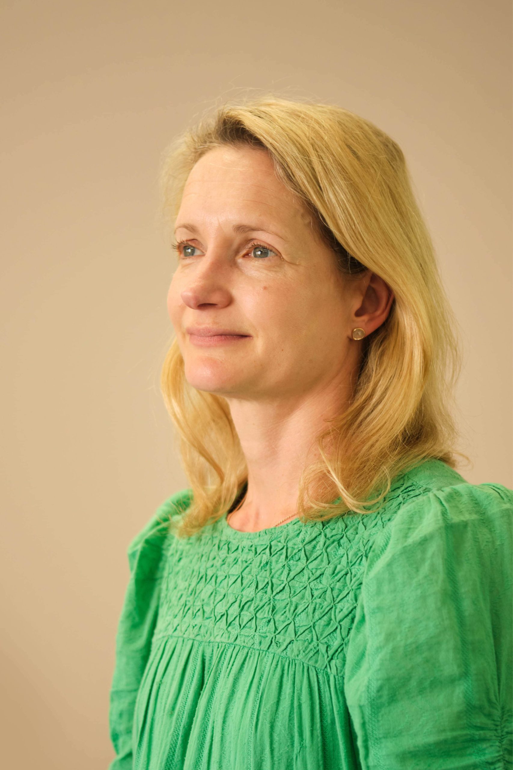 The image shows a person with light blonde hair, wearing a green top. They are looking slightly upward with a neutral expression. The background is a plain light color.