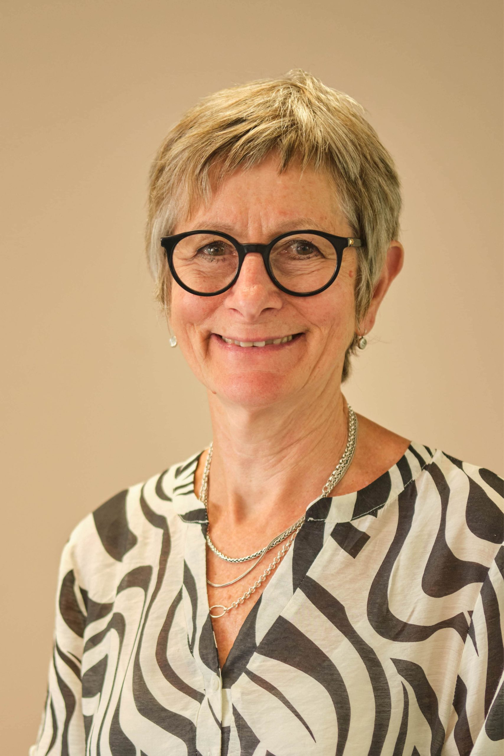 The image shows a person with short gray hair and glasses, smiling. They are wearing a patterned blouse and some necklaces. The background is plain.
