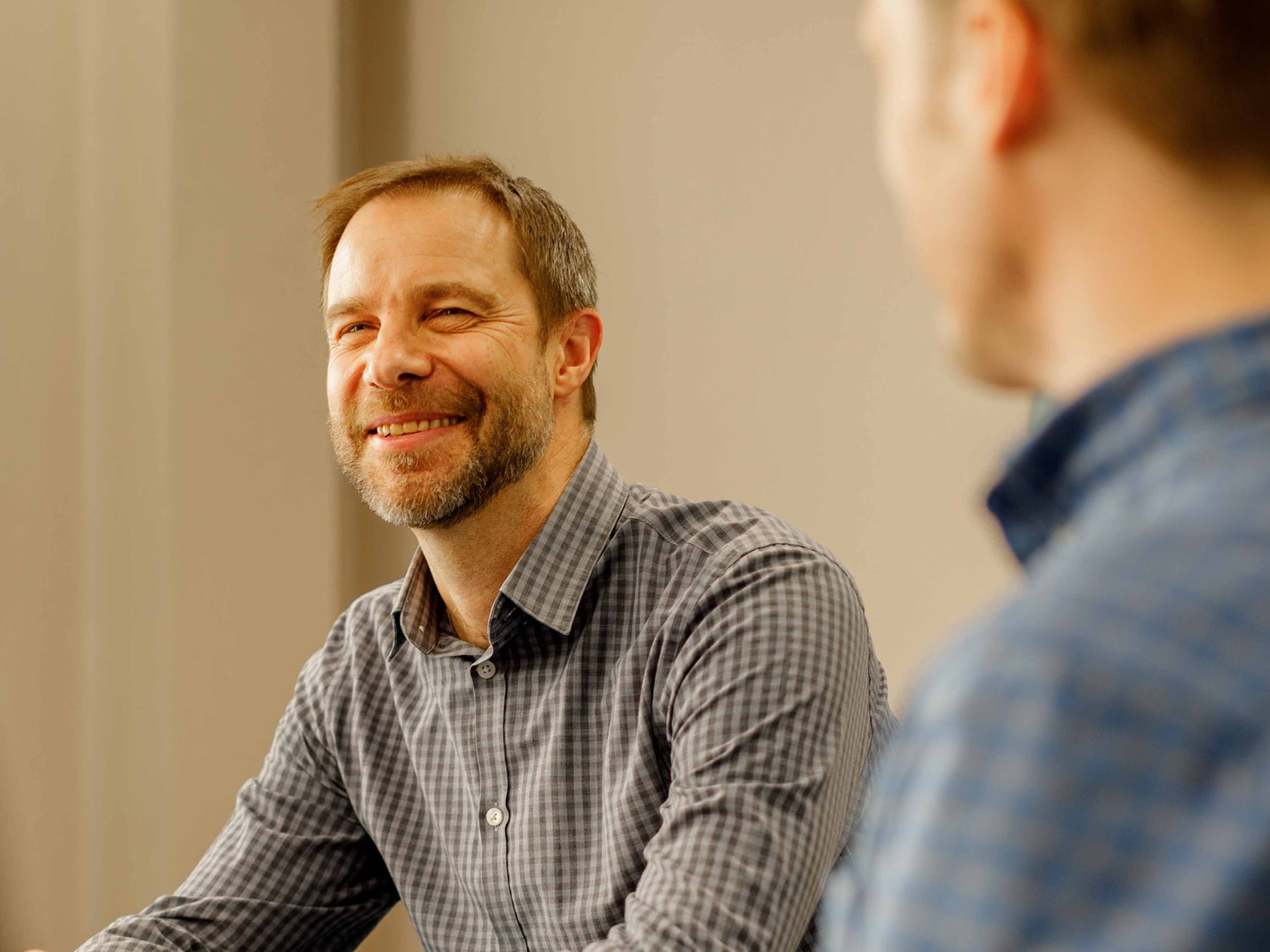 The image shows two people indoors, engaging in conversation. The person in focus is smiling and wearing a checked shirt. The second person is out of focus in the foreground, wearing a blue shirt.