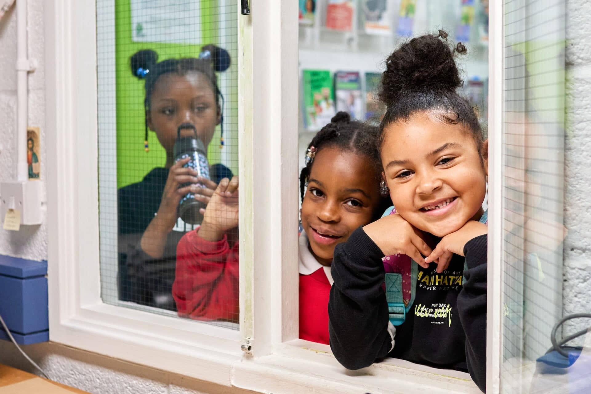 The image shows three children smiling and looking through a window. One child is in the background drinking from a bottle, while the other two are in the foreground, with one wearing a red shirt and the other a black sweater. They seem to be having