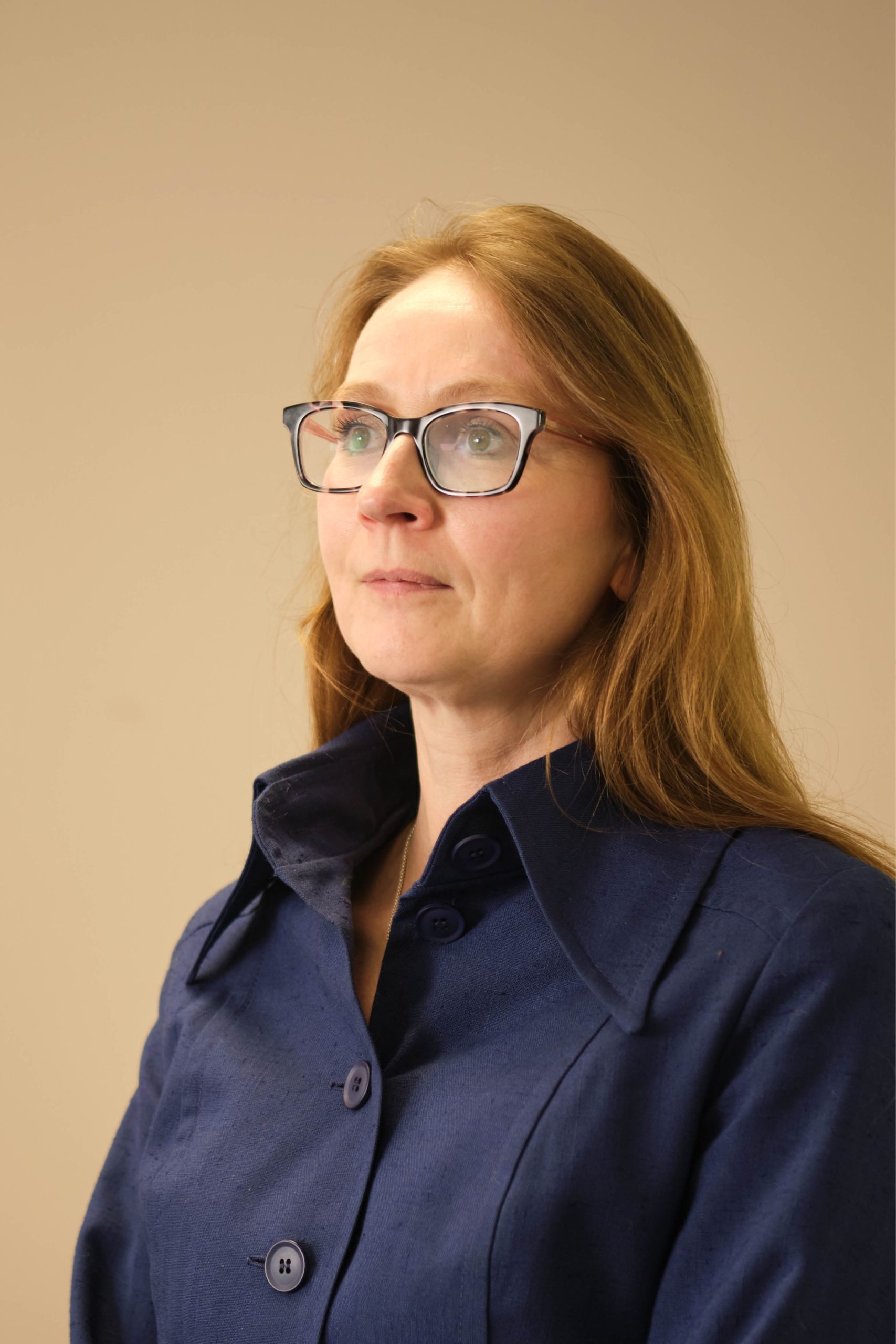 The image shows a woman with glasses and long hair, wearing a blue coat. She is looking slightly to the side against a neutral background.