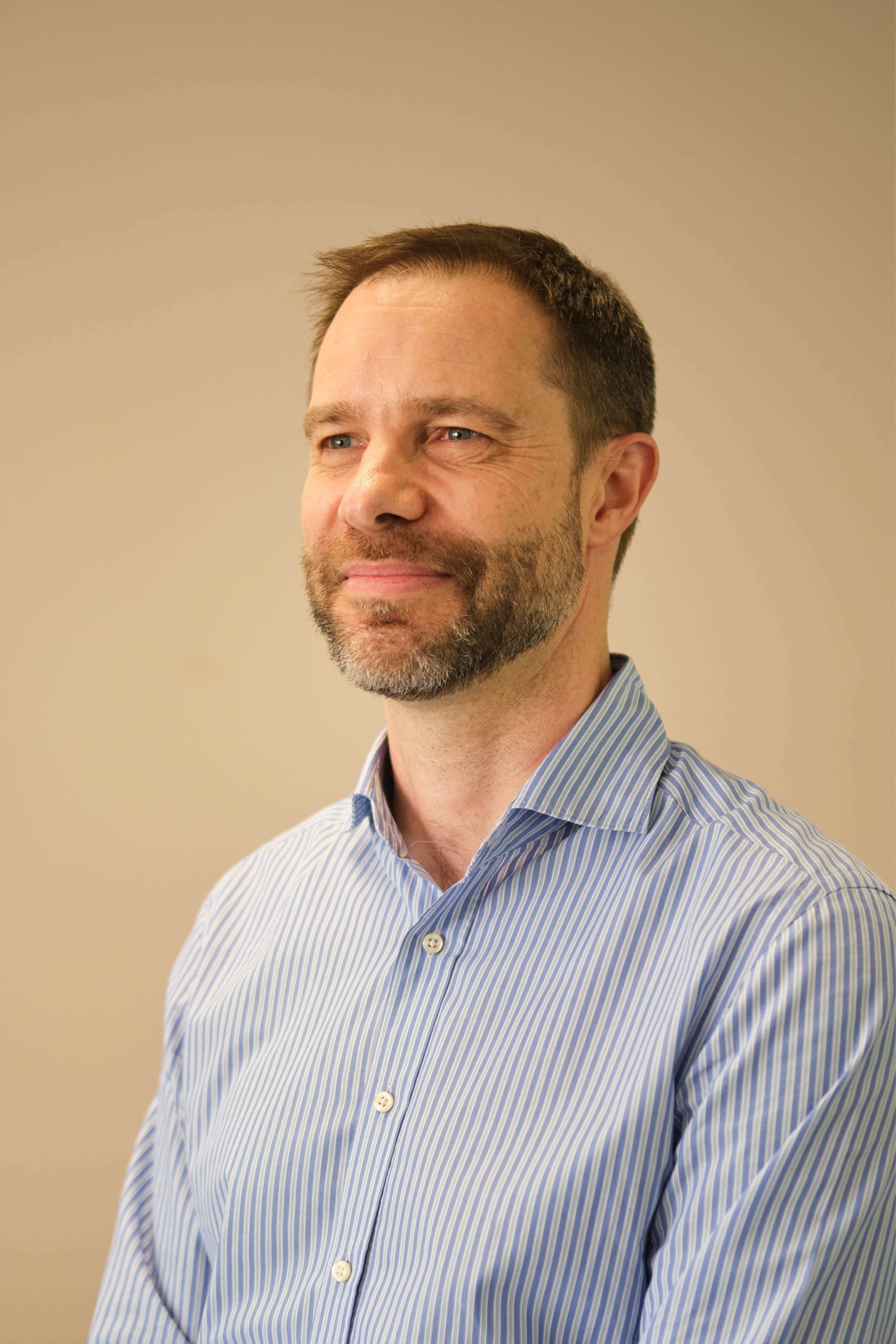 The image shows a person with short hair and a beard, wearing a blue and white striped shirt. The background is plain and neutral in color.