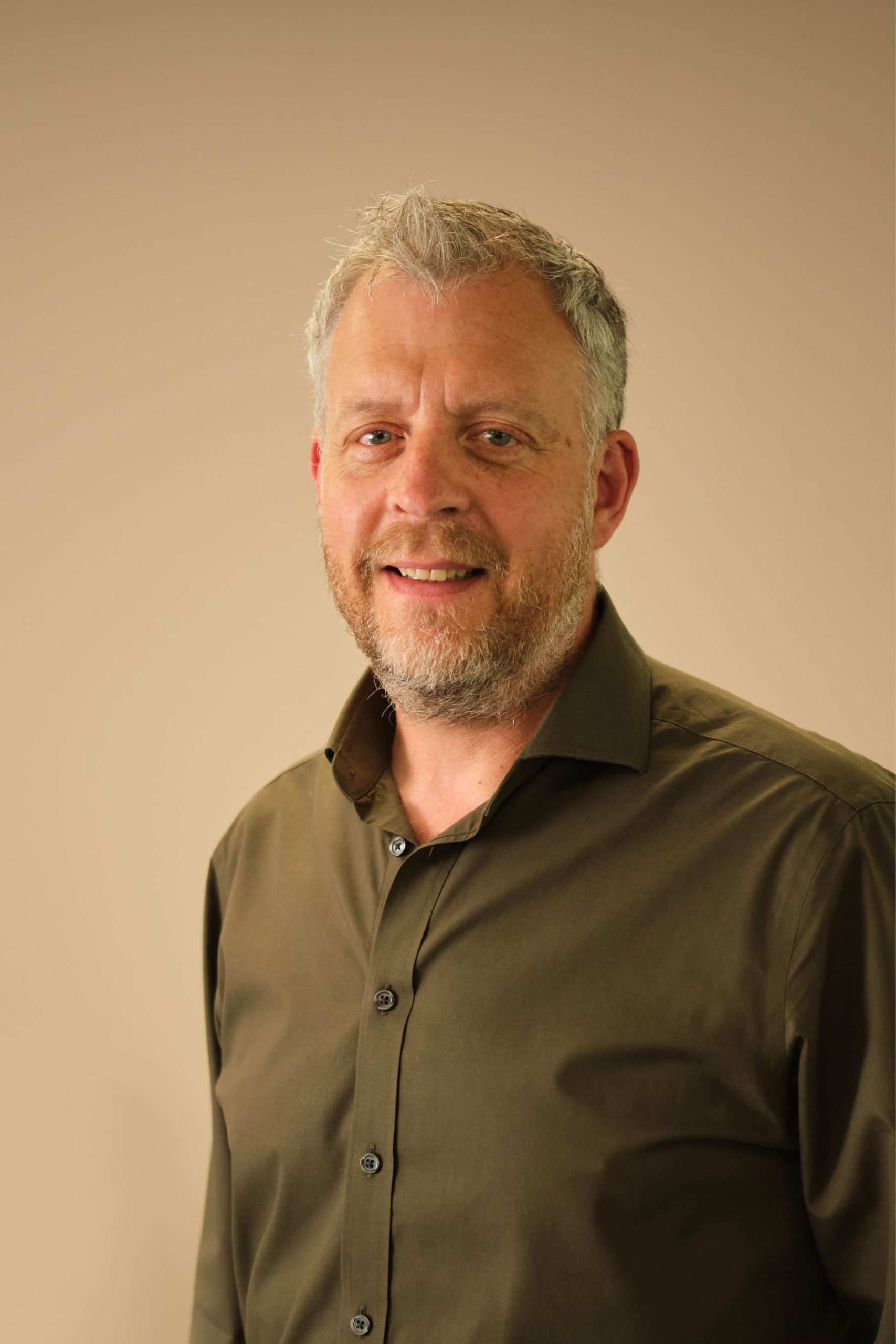 The image shows a man with short hair and a beard, wearing a dark button-up shirt, standing in front of a plain background.