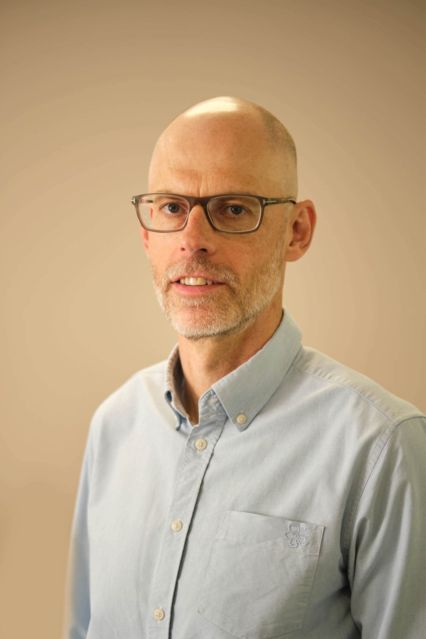 The image is a portrait of a man with a bald head, wearing glasses and a light blue button-up shirt. The background is plain and neutral-colored.