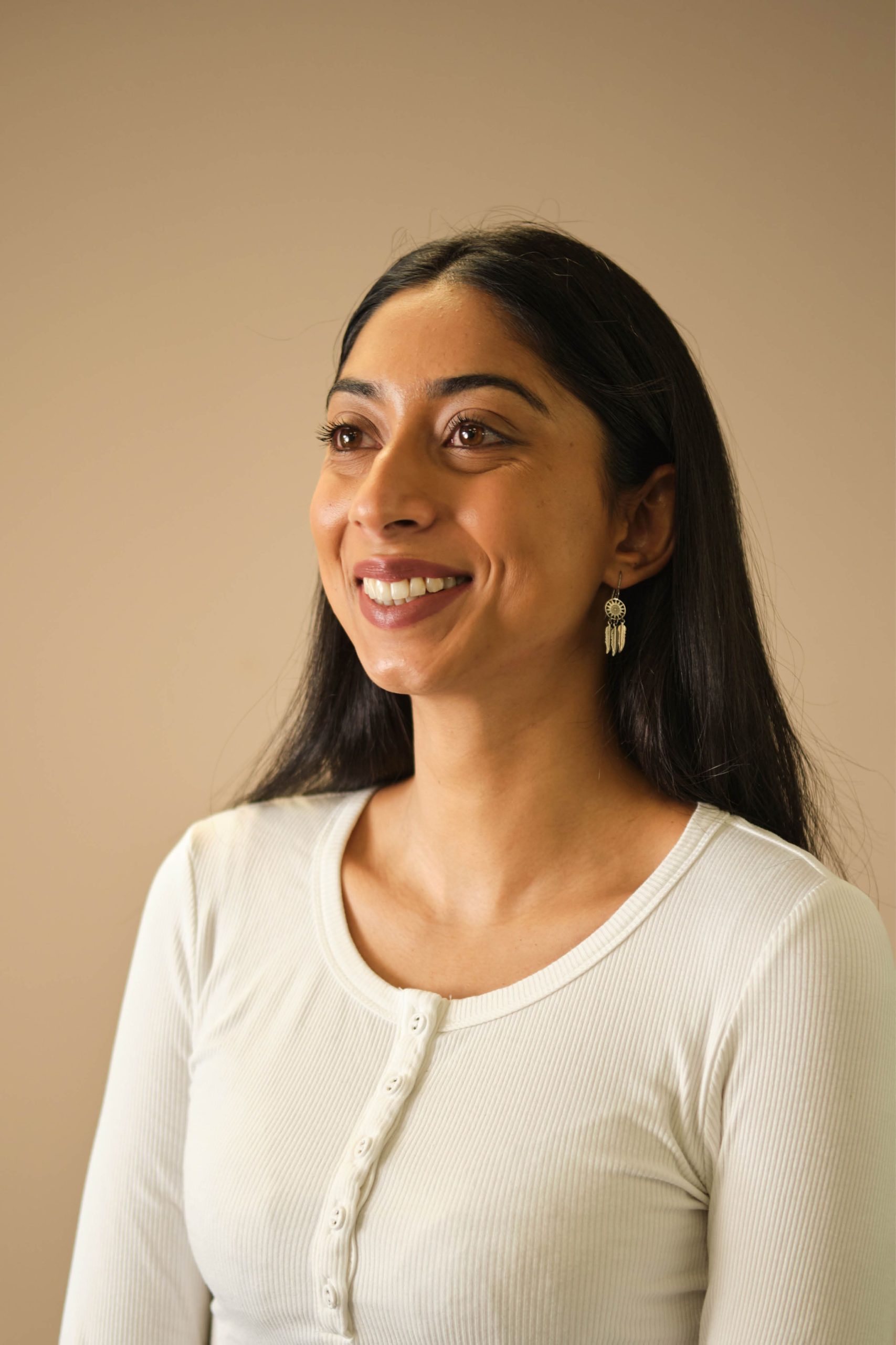 The image shows a woman with long dark hair, wearing a white ribbed shirt with buttons. She has earrings and is smiling, looking slightly to the side. The background is plain and neutral-colored.