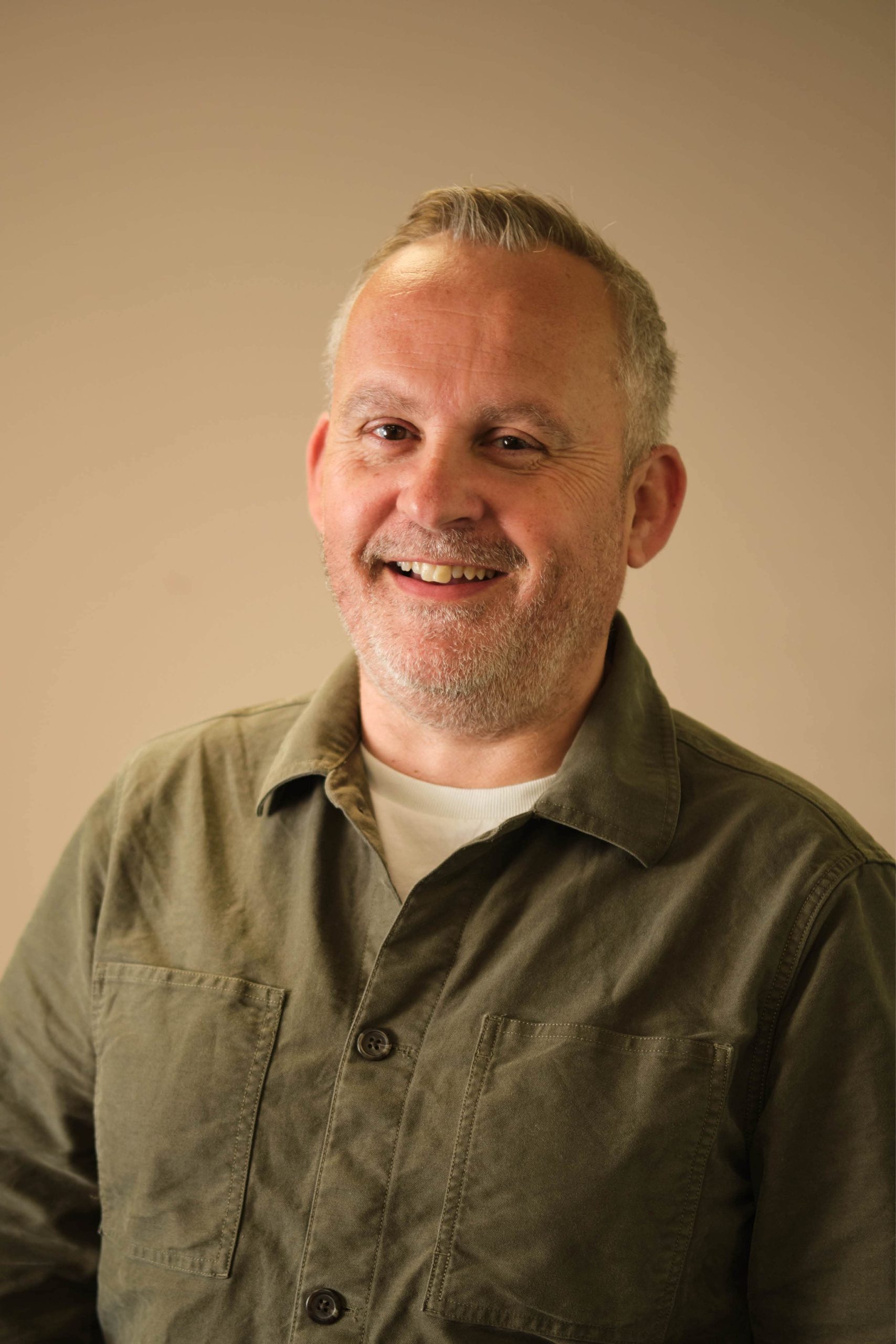 The image shows a person smiling and wearing a green shirt over a white undershirt. The background is plain and beige.