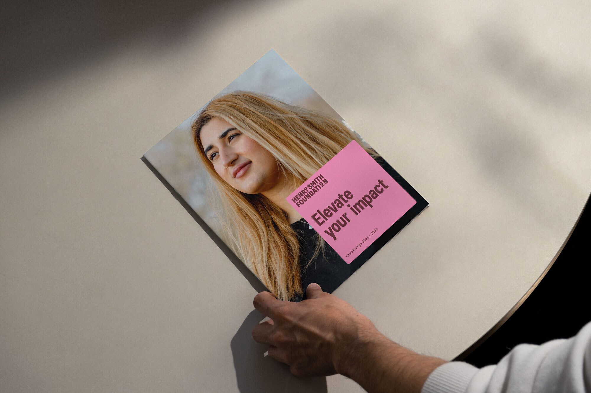 The image shows a person holding a brochure or booklet with a cover that features a portrait of a person with long hair. The text on a pink background says HENRY SMITH FOUNDATION. Elevate your impact. Our strategy 2023-