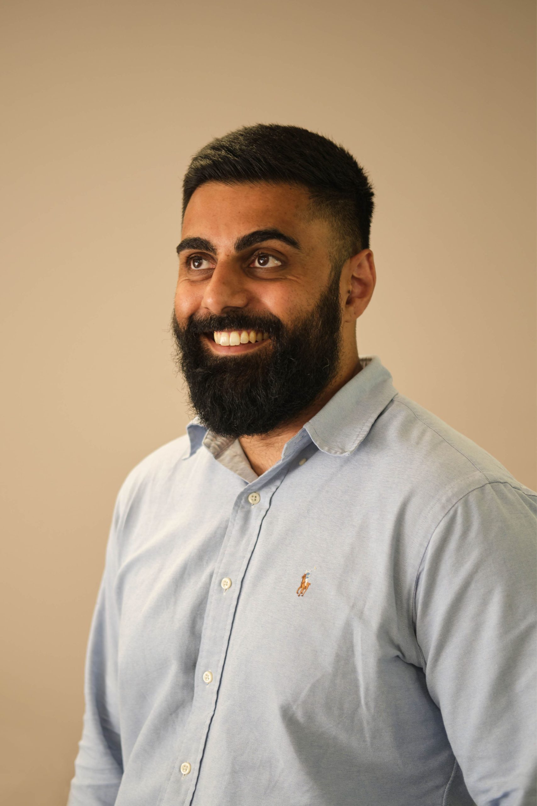 The image shows a person with a beard and short hair, wearing a light blue button-up shirt. The background is plain and neutral-colored. The person is smiling.