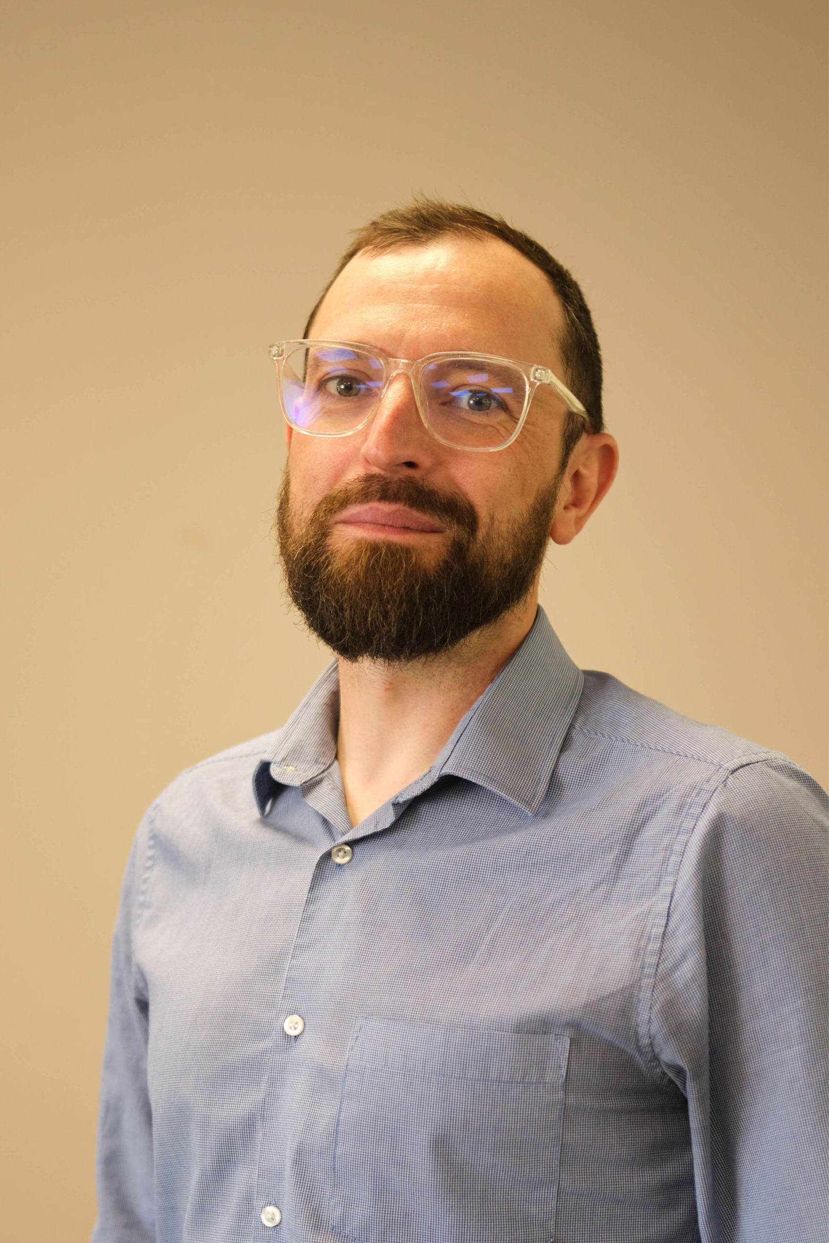The image shows a person wearing a light blue button-up shirt and clear glasses. They have a beard and are standing against a plain background.