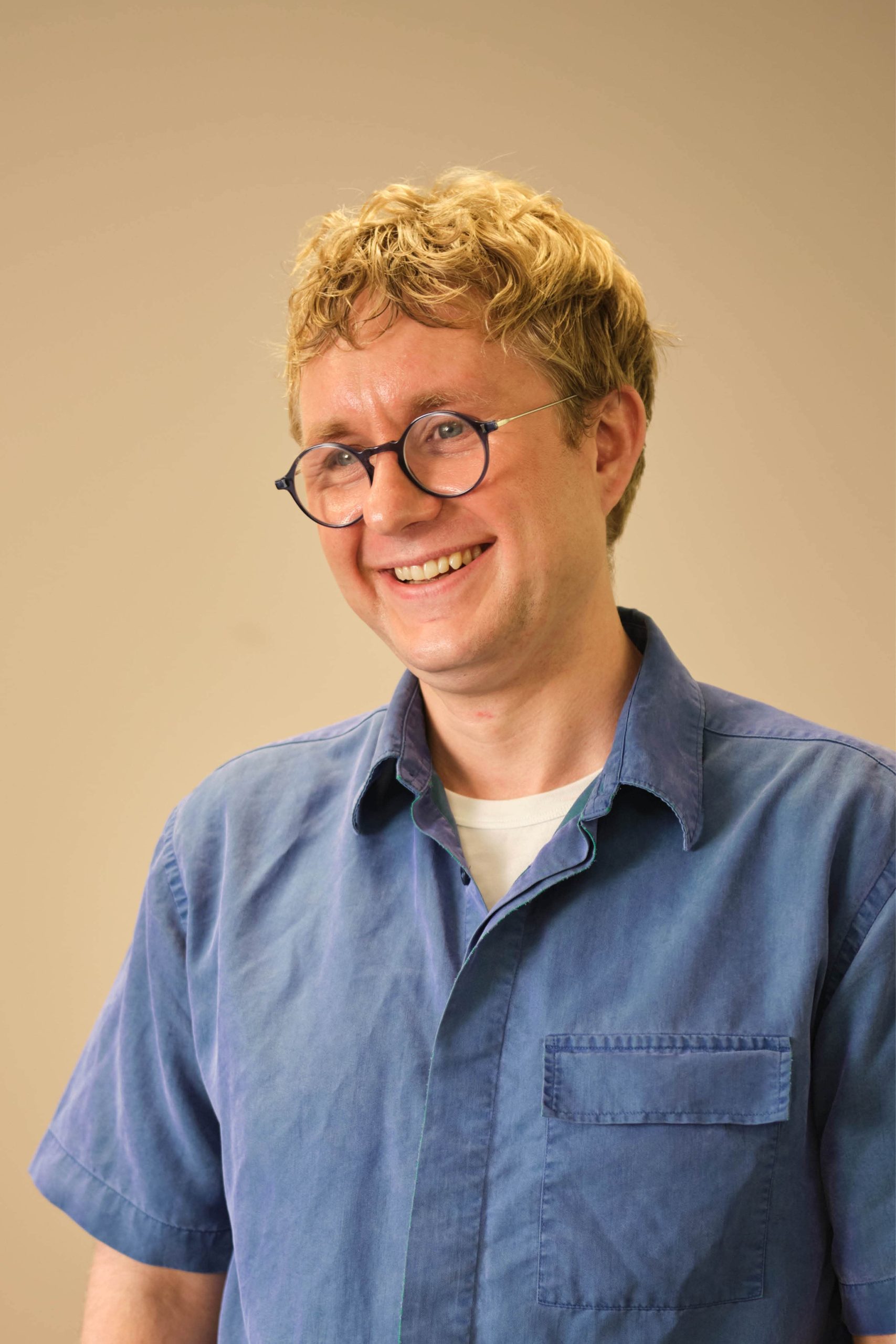 The image shows a person with blond hair and glasses, smiling. They are wearing a blue shirt over a white undershirt. The background is plain and neutral in color.