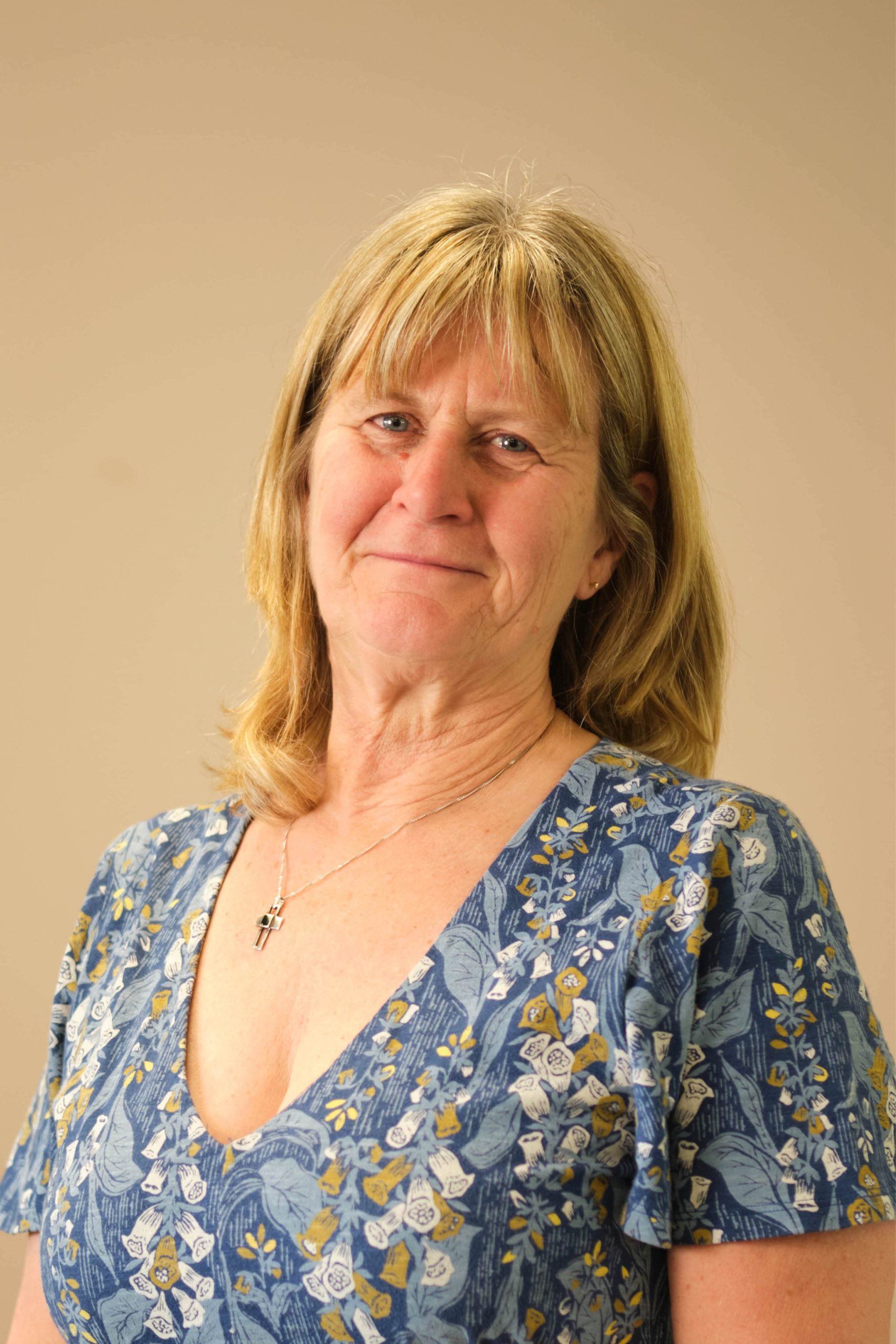 The image shows a person with shoulder-length hair, wearing a blue floral-patterned top and a necklace with a cross pendant. The background is a plain, neutral color.