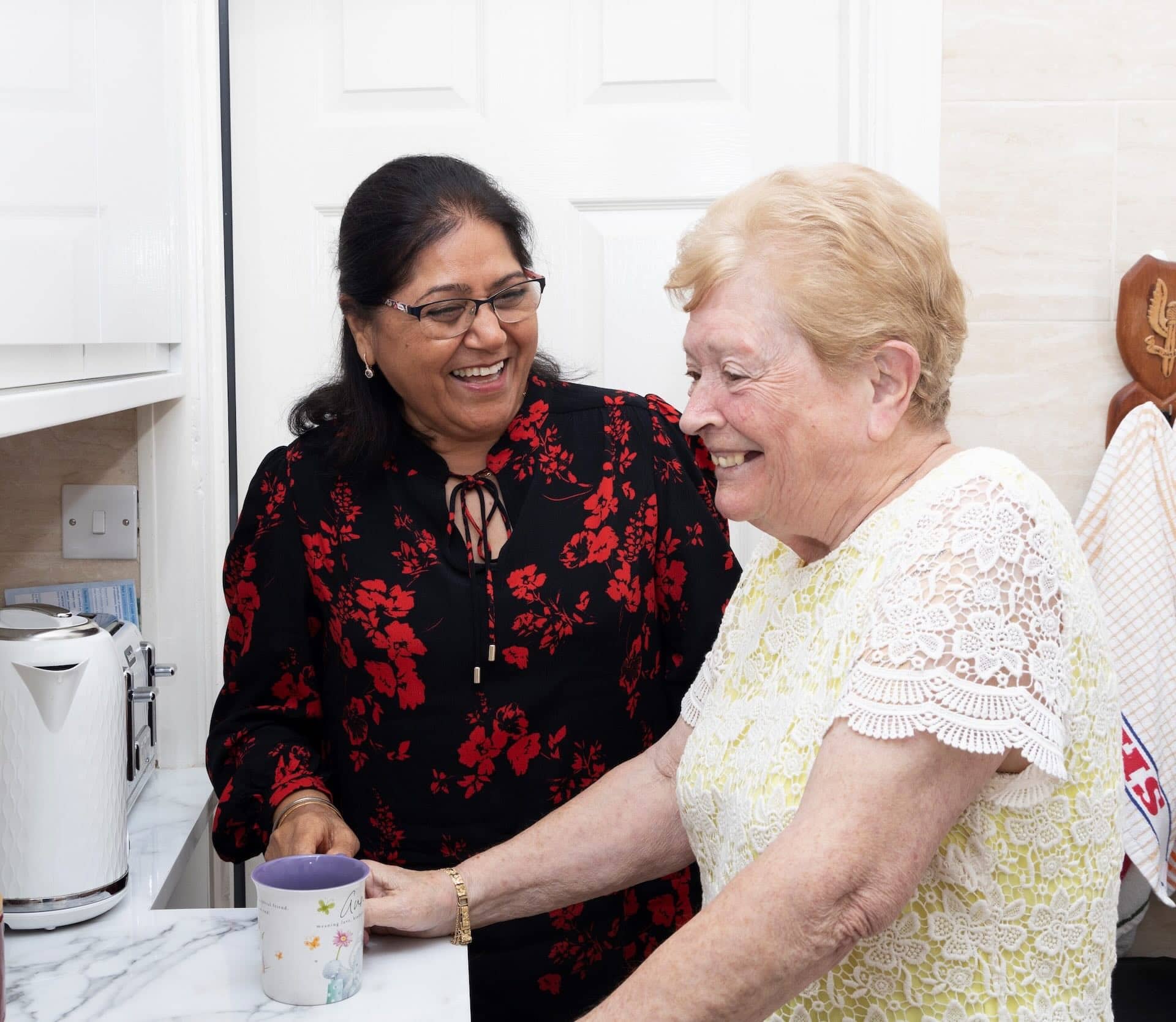 The image shows two women in a kitchen, smiling and engaged in conversation. One woman is holding a mug. There is a kettle and some appliances on the countertop.