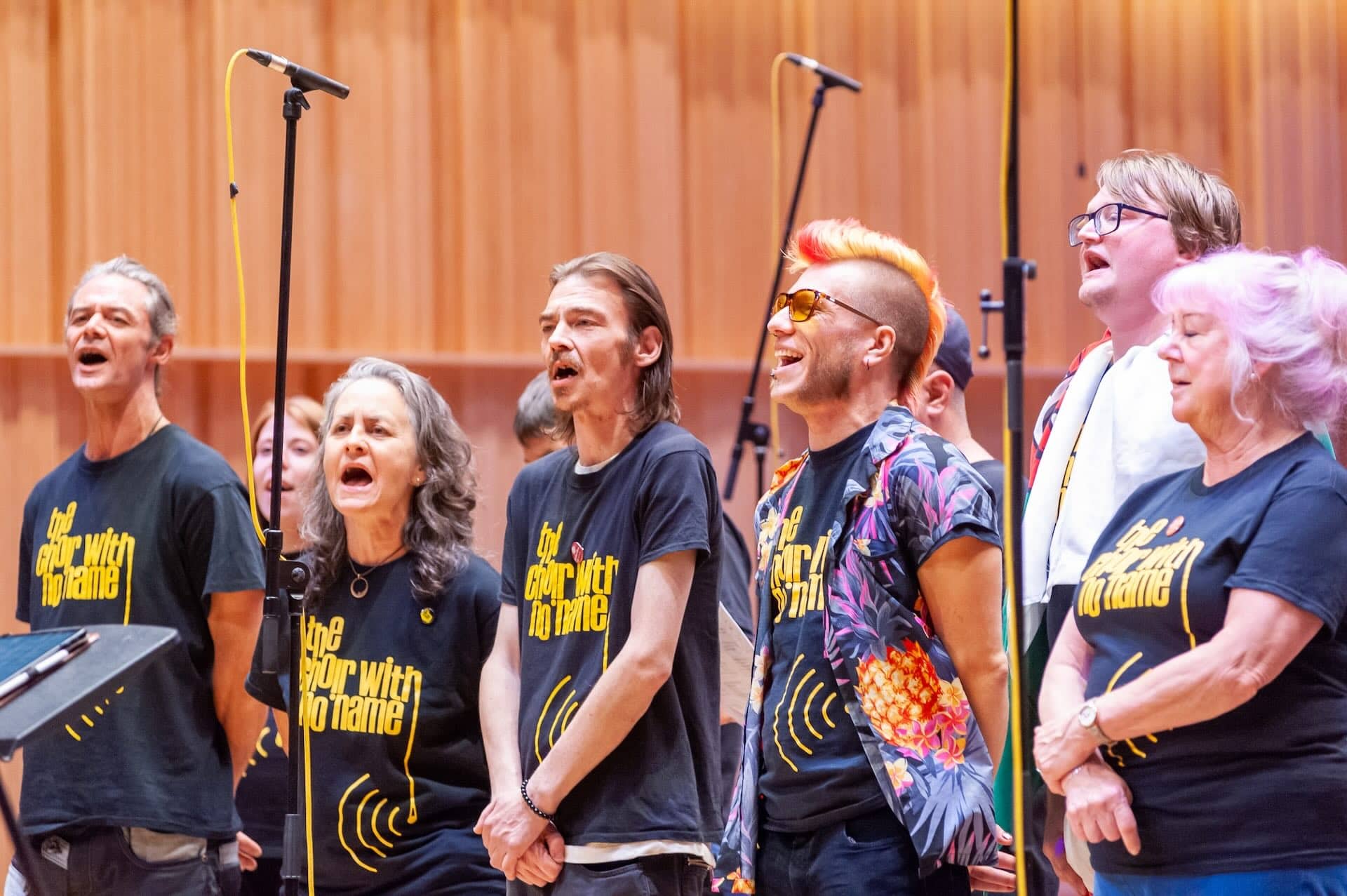 The image shows a group of people singing, likely as part of a choir. They're standing in front of microphones and are wearing matching t-shirts that read The Choir with No Name. The setting appears to be indoors, possibly in a concert hall or