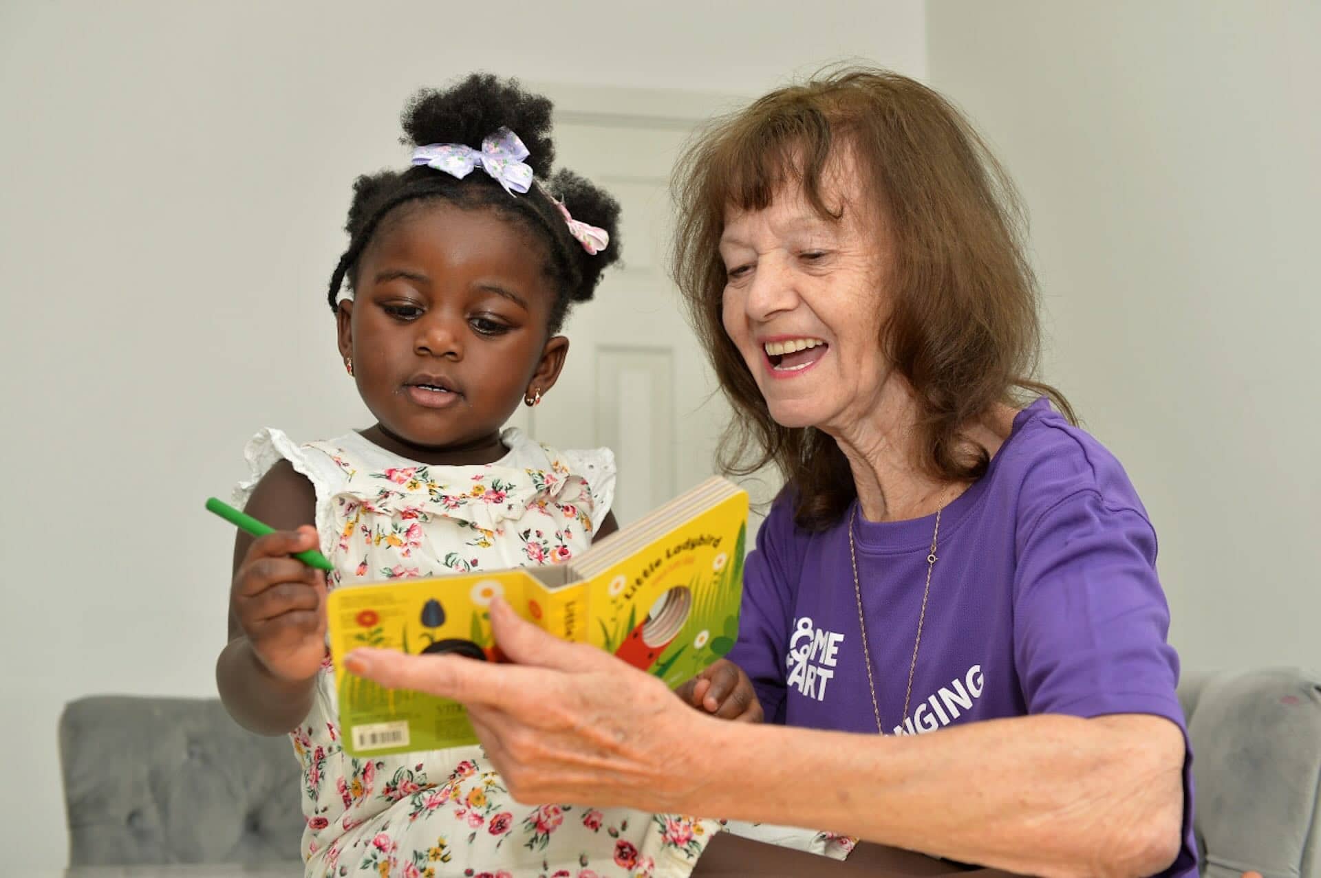 The image shows a young child and an older adult reading a book together. The child is holding a green crayon, and the older person is smiling as they look at the book. The older adult is wearing a purple shirt with white text. The