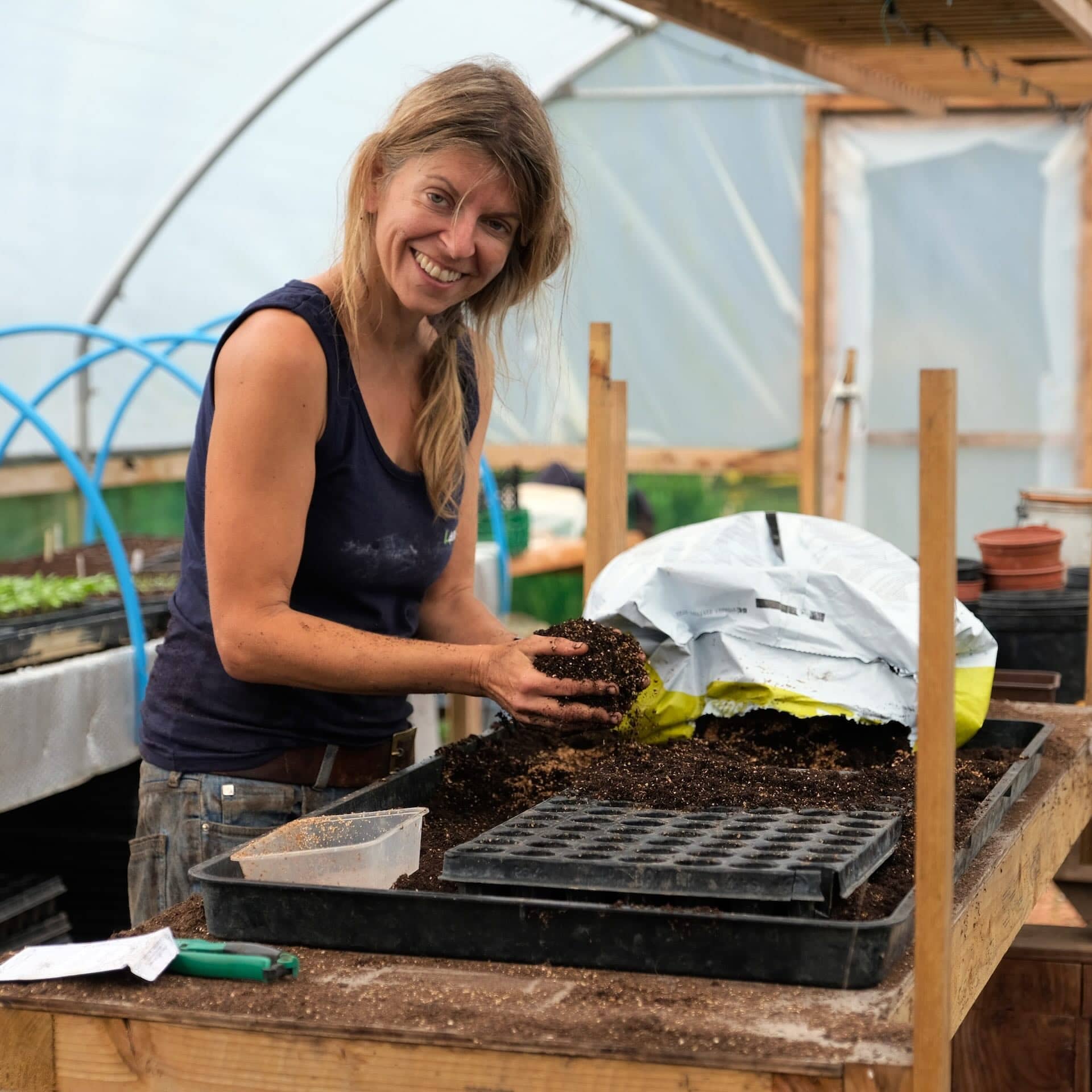 The image shows a person in a greenhouse working with soil. They appear to be setting up seed trays or preparing soil for planting, with a bag of soil nearby. The environment looks like a gardening workspace, possibly for propagating plants or gardening.
