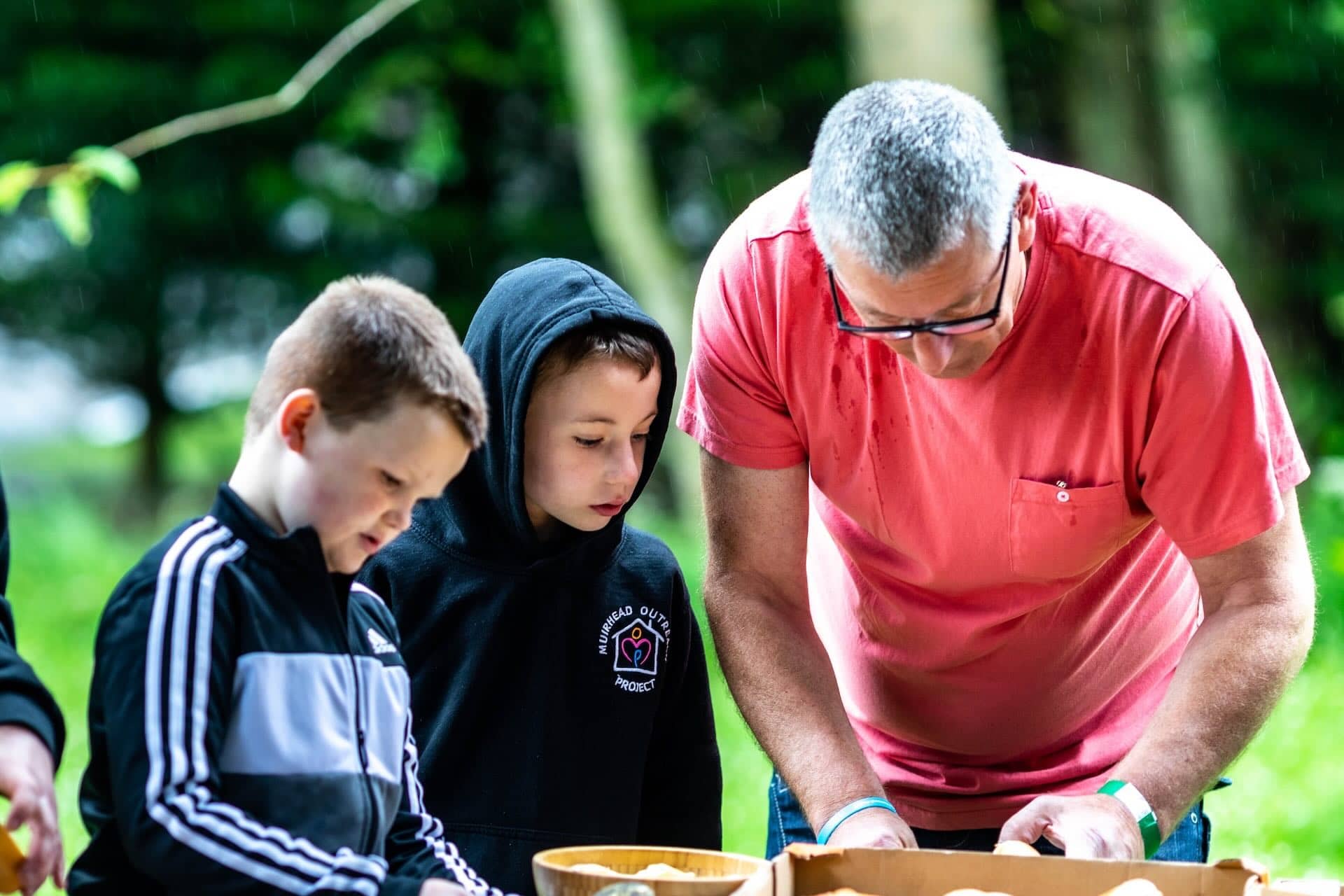 The image shows three people outdoors engaged in an activity together. Two are children and one is an adult. They're looking at something within a wooden container. The scene is set in a forested area, as evident from the blurred greenery in the background.