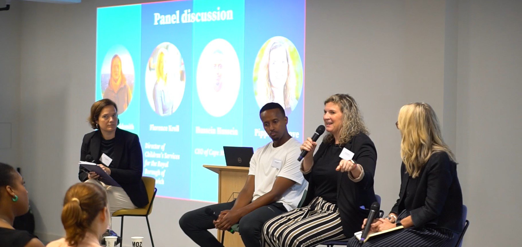 The image shows a panel discussion taking place. There are four people seated with microphones, indicating they are part of the panel. A screen in the background displays text and pictures related to the discussion, although it's not entirely clear from this angle. The setting
